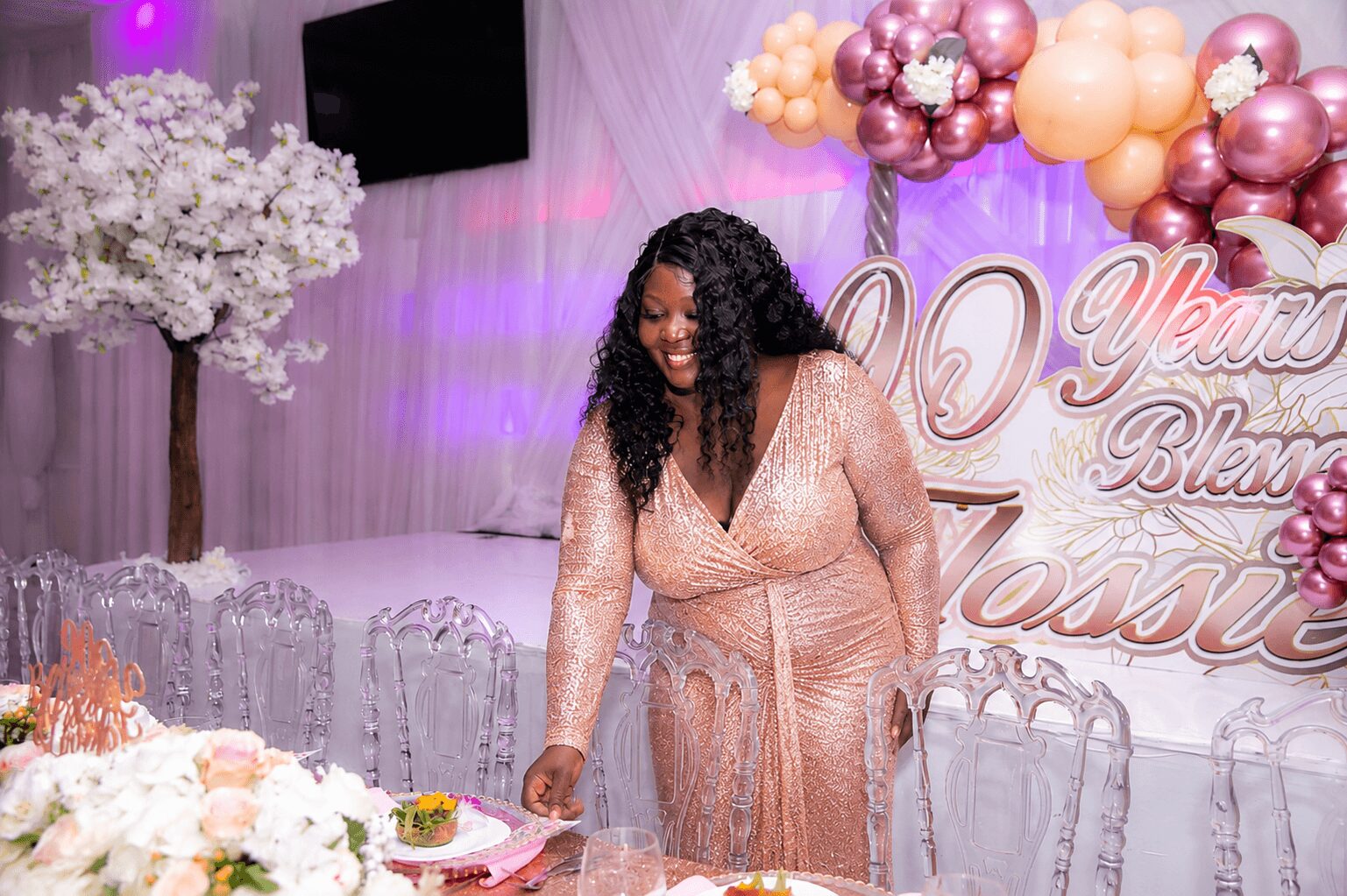 Woman in a pink dress at celebration with balloons and floral decorations, smiling and reaching for a plate.