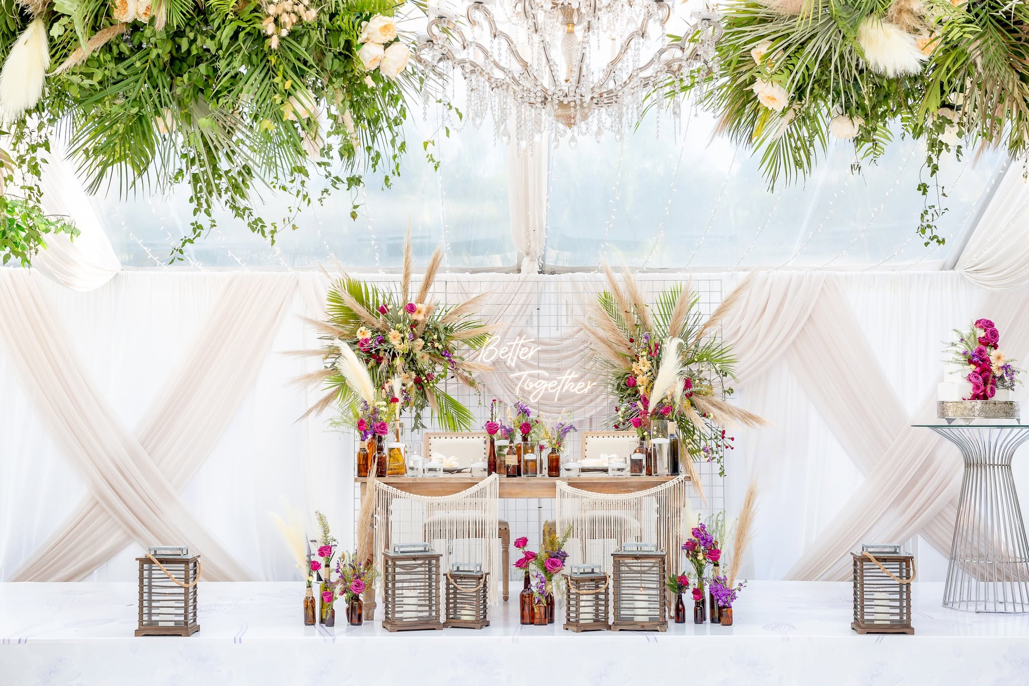 Decorated table with floral arrangements and candles, set against a white draped backdrop with hanging greenery and flowers above.