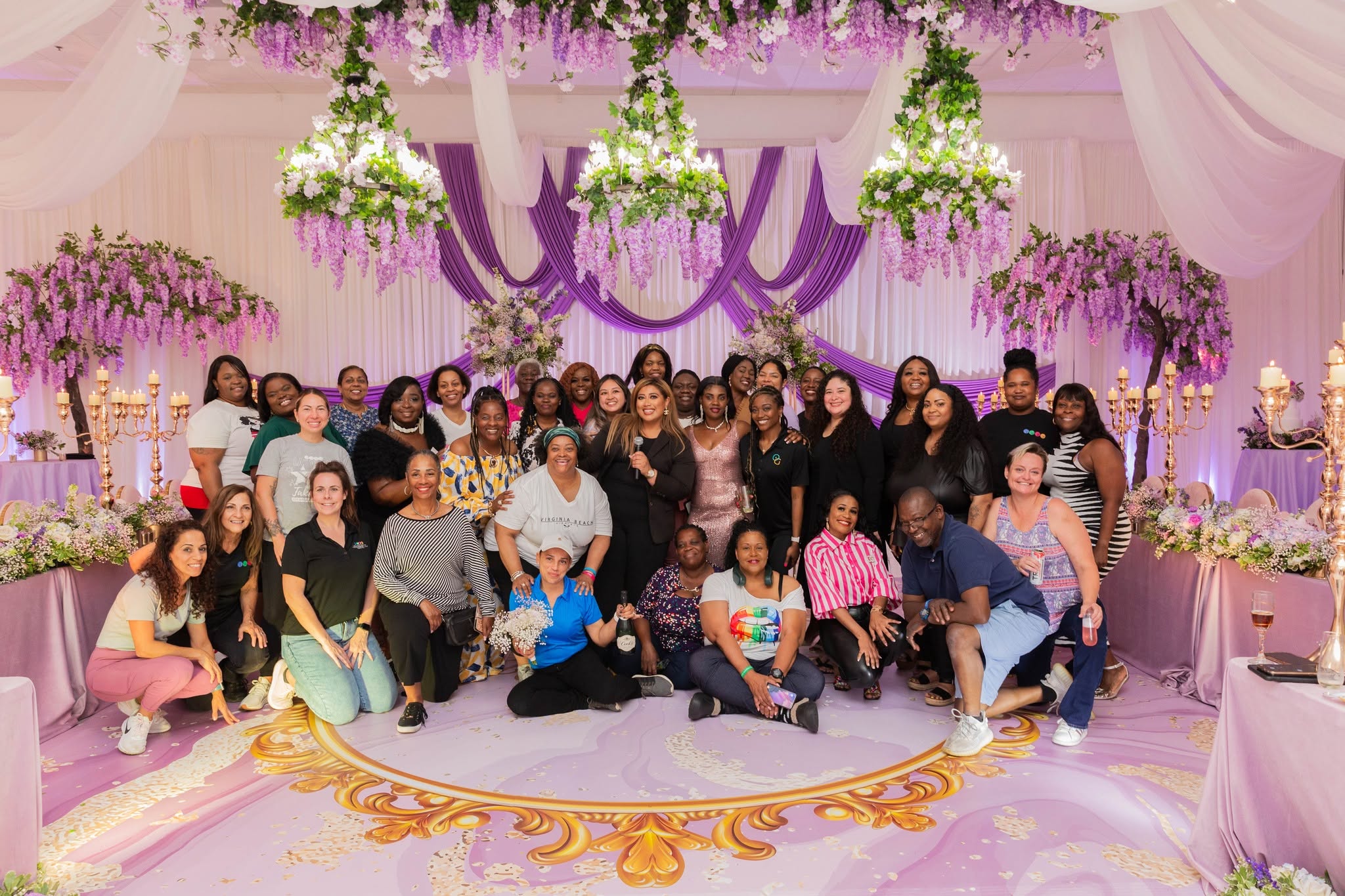 Group of people gathered in a decorated event space with purple and white drapery, floral arrangements, and hanging lights.