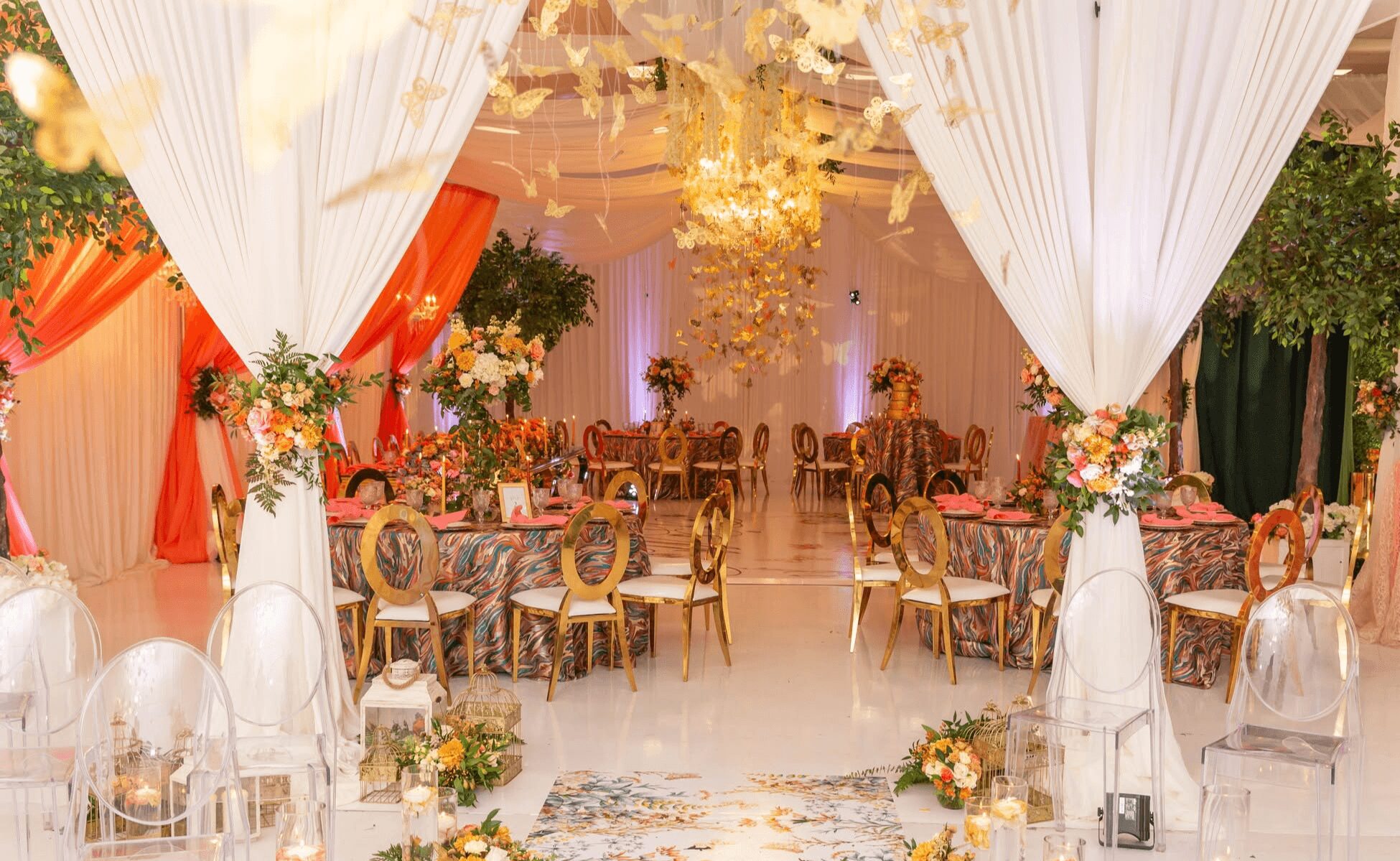 Elegant decorated event space with draped white curtains, floral arrangements, and ornate chairs, illuminated by a chandelier, with a stage in the background.