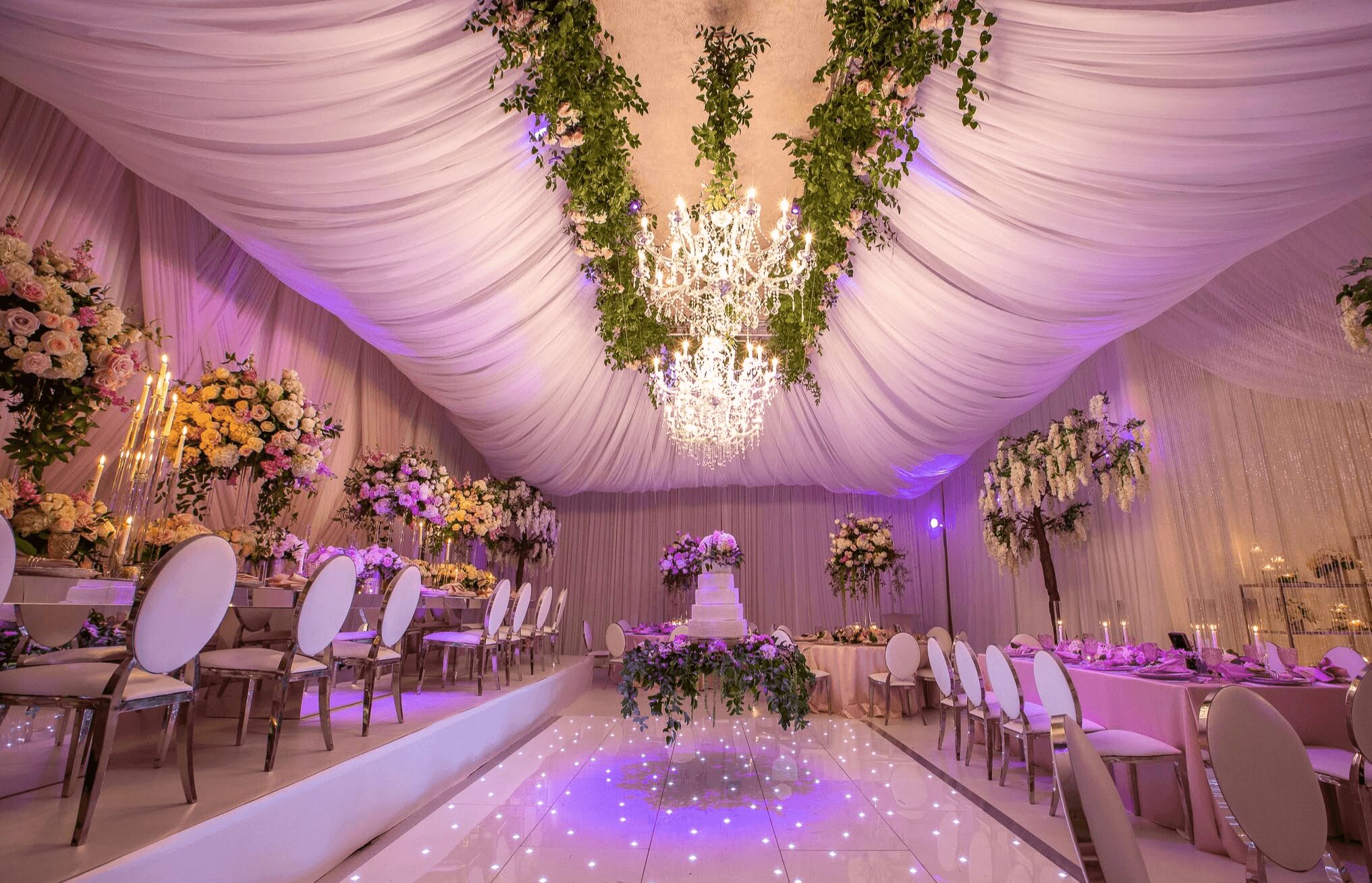 Elegant decorated event space with draped ceiling, chandelier, floral arrangements, and a dance floor, set for a celebration.