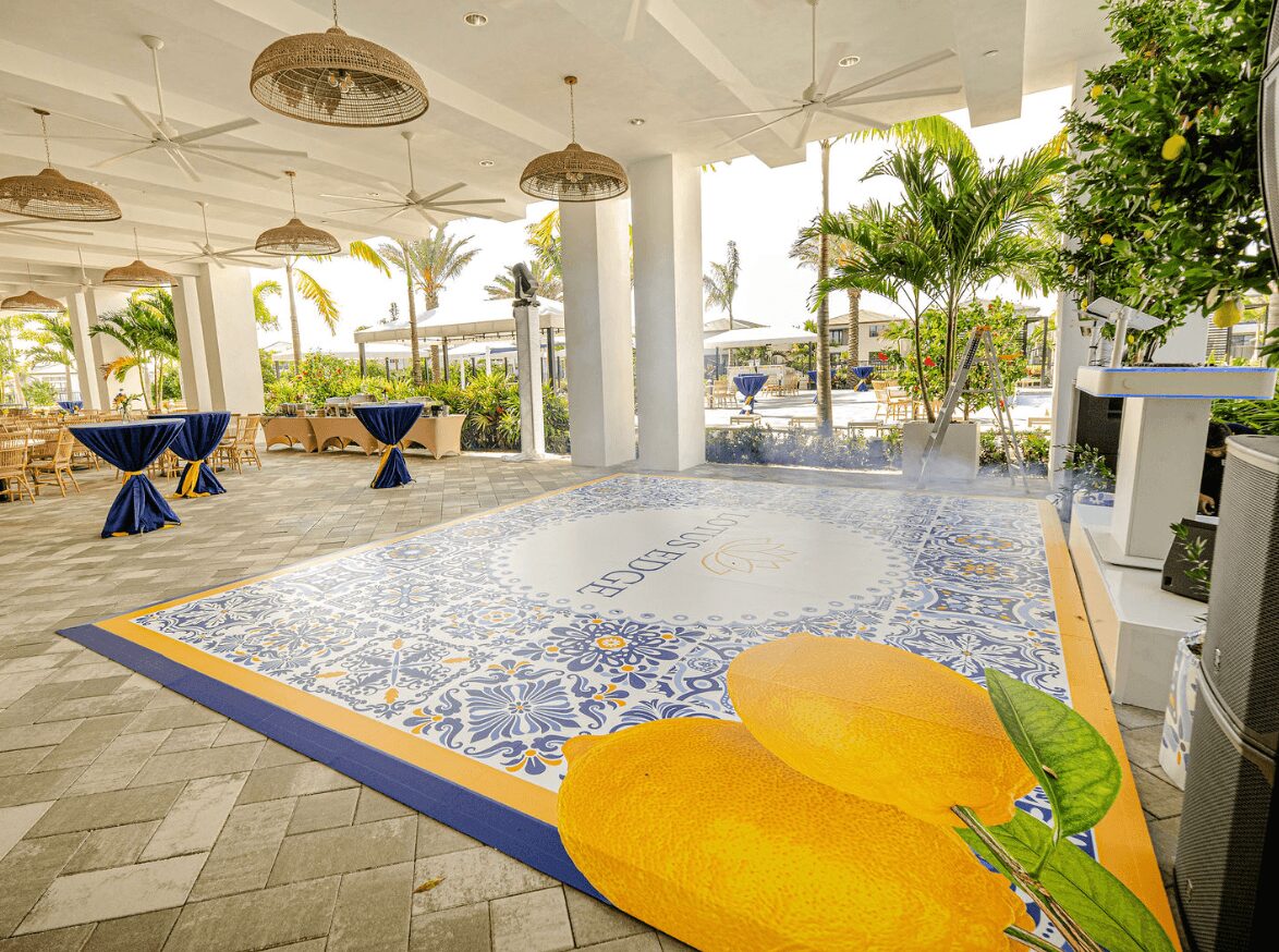 Indoor pool area with decorative tile floor, tables with blue tablecloths, and tropical plants outside. Yellow lounge chairs in foreground.