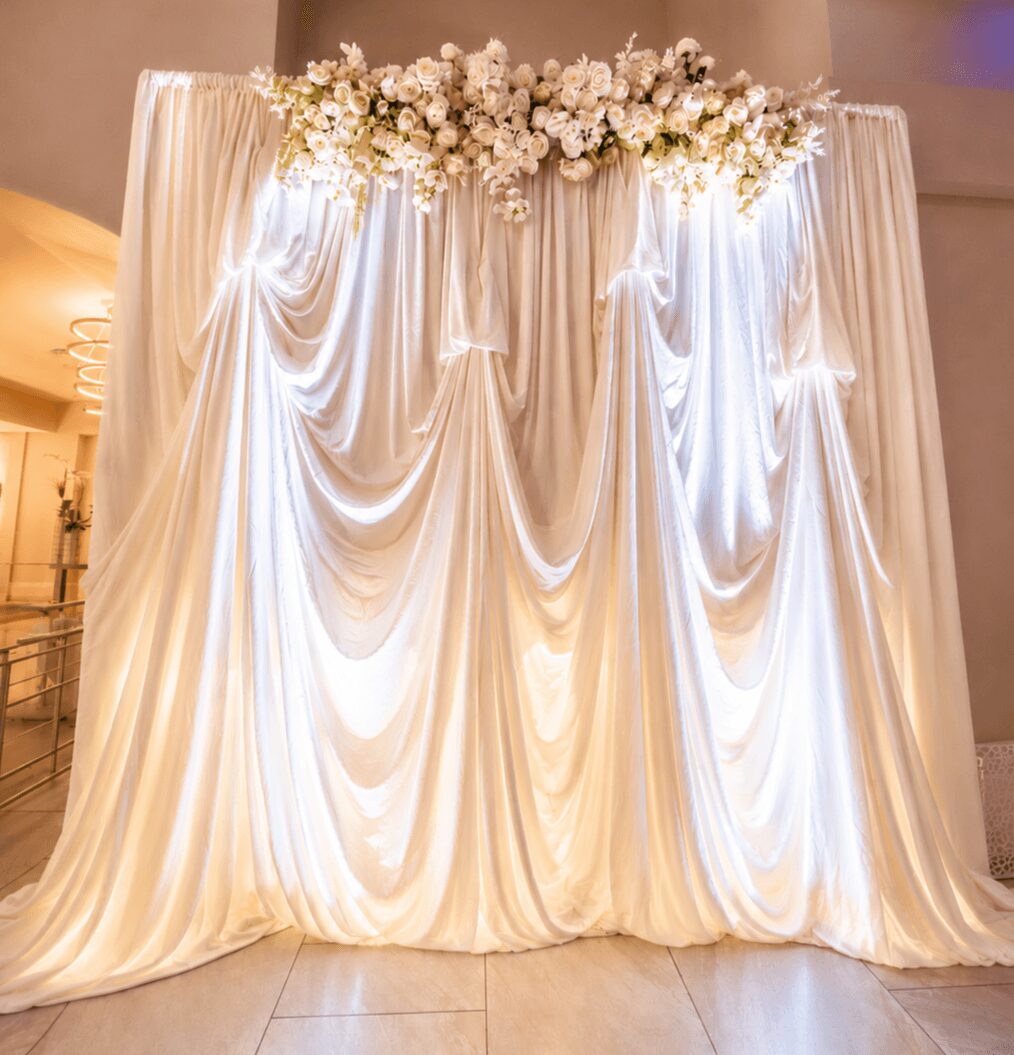 Elegant white draped curtain backdrop with floral arrangement at the top, used for a special event or wedding.