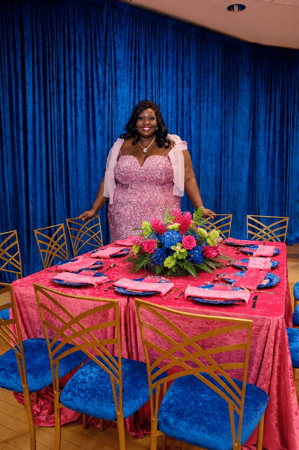 Woman in pink dress standing behind a decorated table with a floral centerpiece, blue curtains in background.