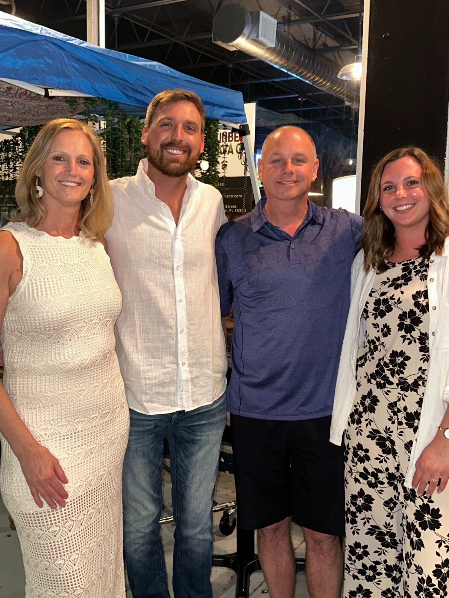 Four people standing together outdoors, smiling, with a blue canopy and string lights in the background.