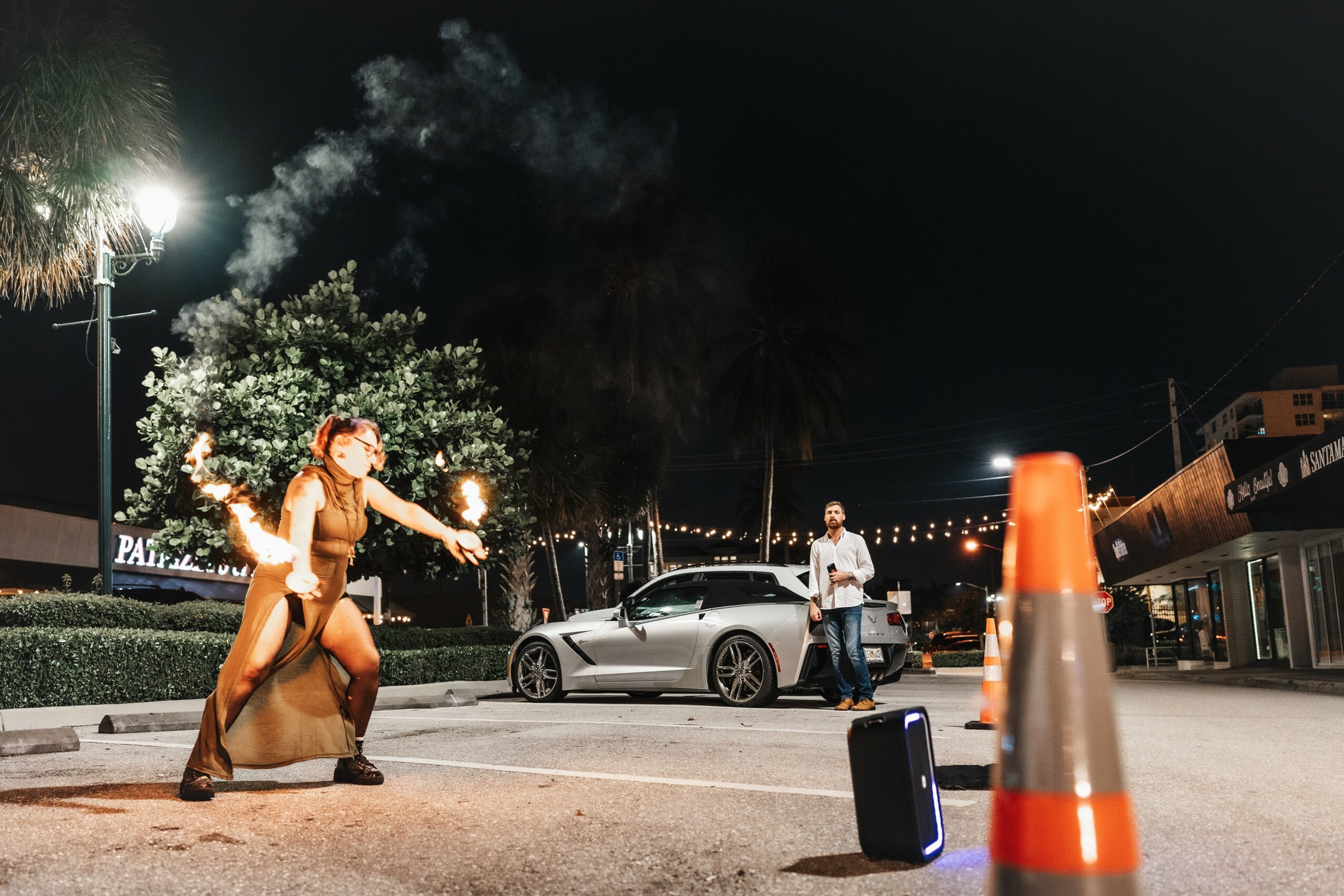 Person performing fire spinning on street at night with onlookers, car, and traffic cone nearby.