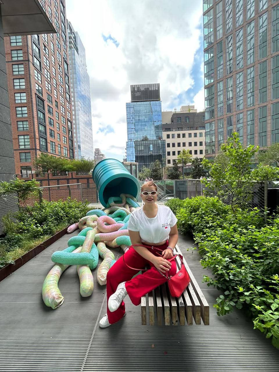 Woman sitting on a bench in a city park with tall buildings in the background, greenery, and a colorful sculpture, wearing sunglasses and a white top.
