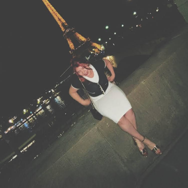 Person standing on a sidewalk at night with the Eiffel Tower illuminated in the background.