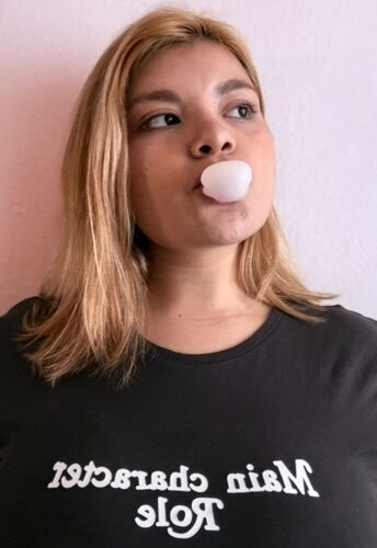 Young woman with blonde hair blowing bubble gum, wearing a black shirt with white text, against a light background.