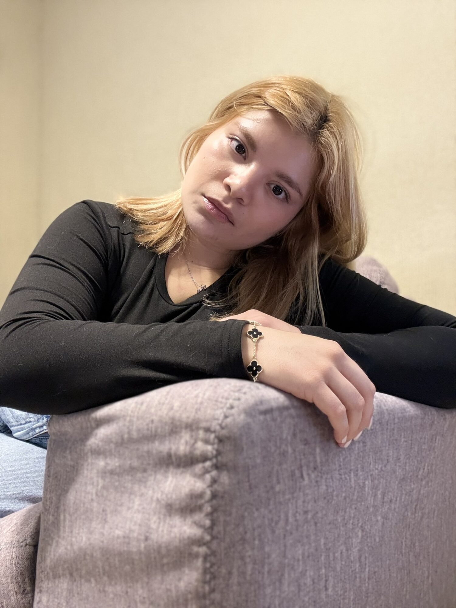 Young woman with blonde hair resting her arm on a gray sofa, looking at the camera, neutral expression, beige background.