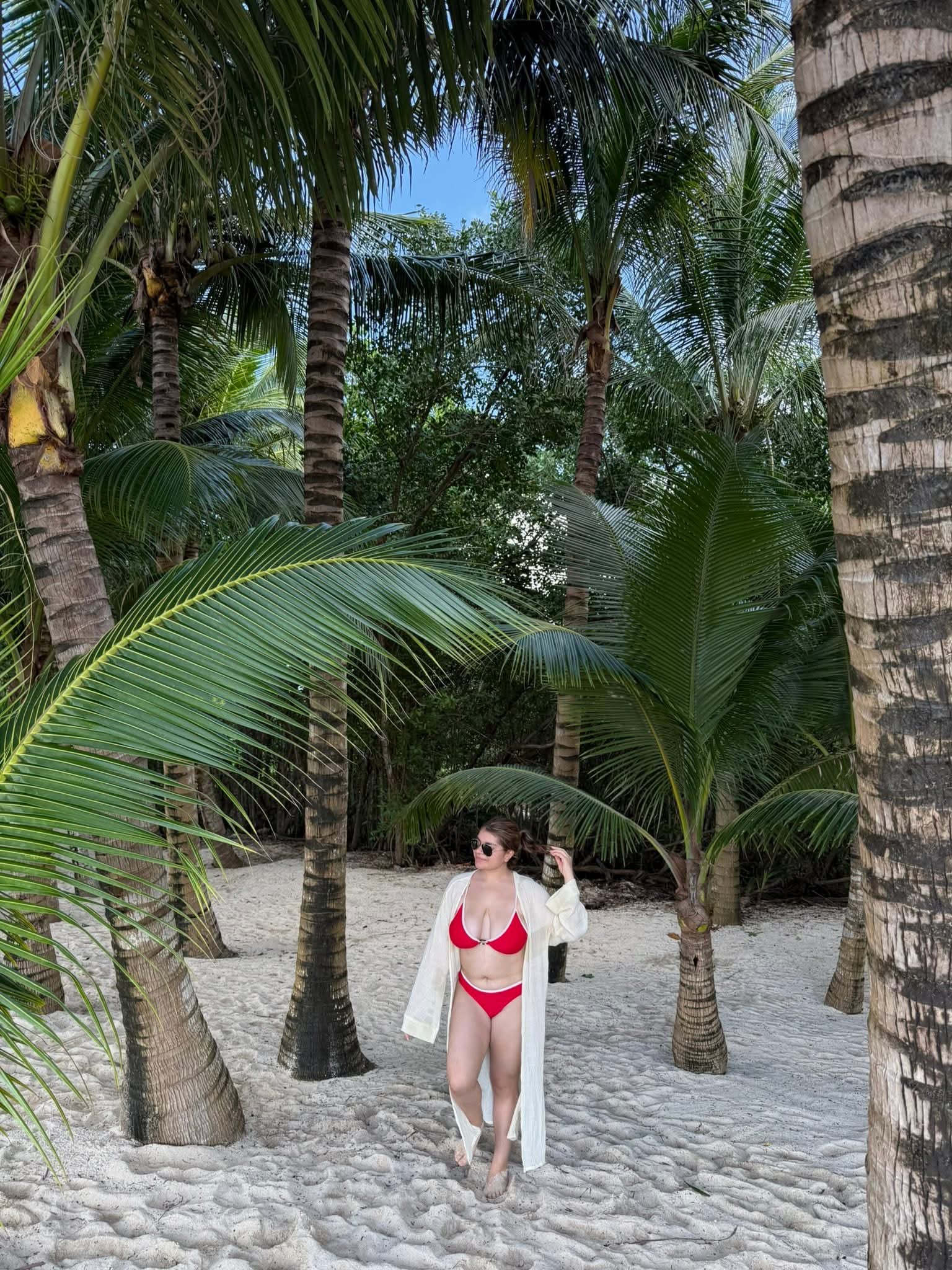 Woman in red swimsuit and white cover-up walking on sandy beach surrounded by palm trees.