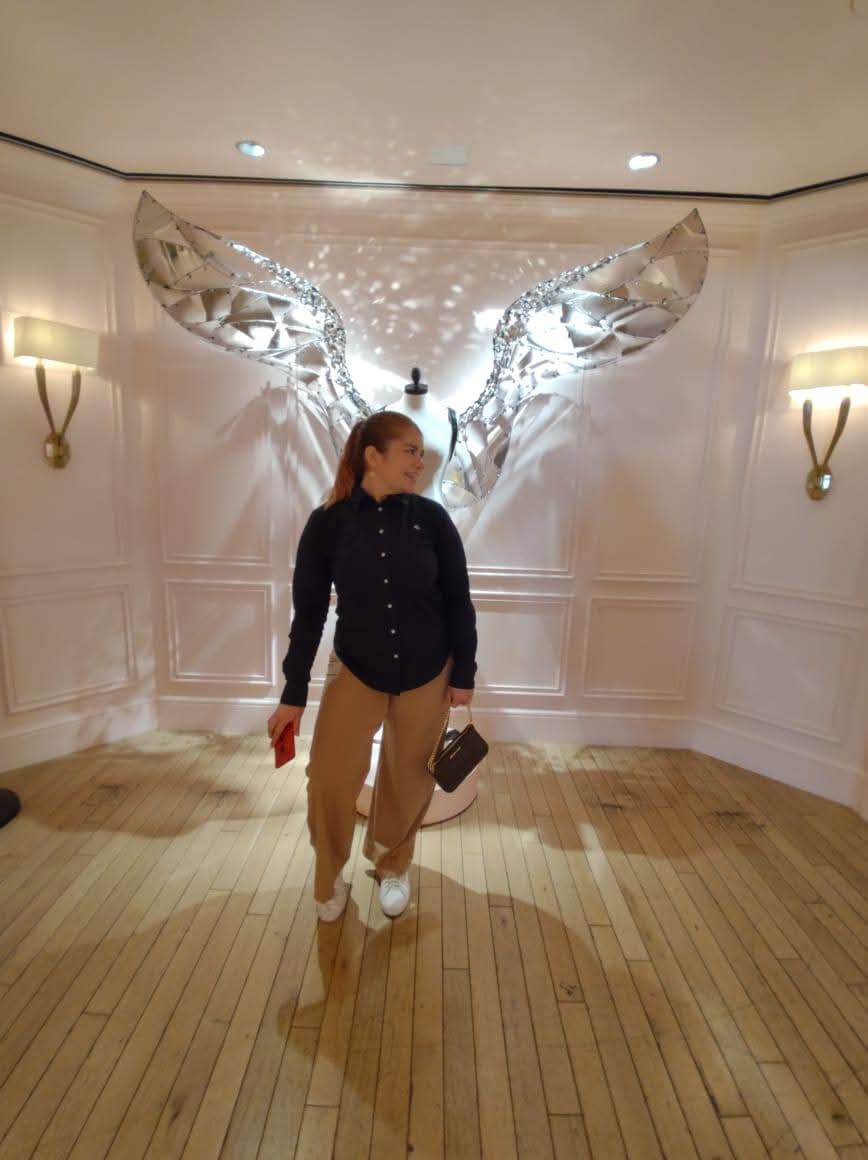 Person standing in front of illuminated angel wing display, holding a hat, in a room with wooden floor and wall sconces.