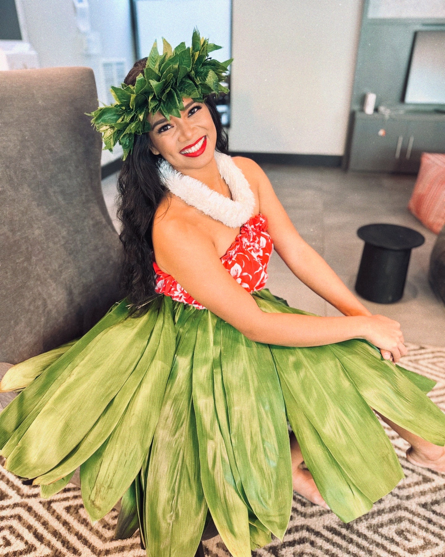 Woman in traditional dress with green leaf crown, smiling, sitting on floor in modern room