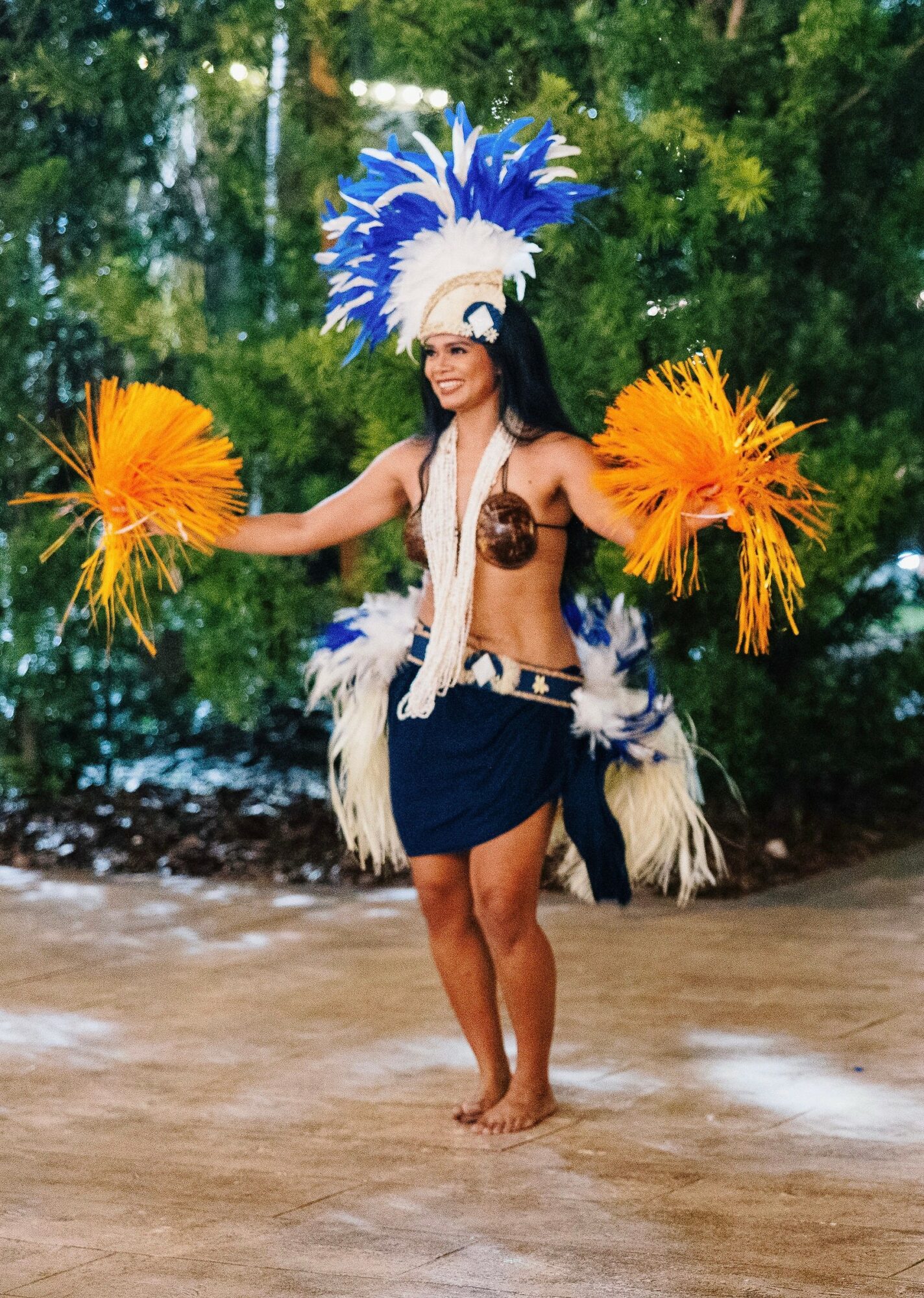 Woman in traditional costume with feathered headdress and yellow pom-poms dancing outdoors, surrounded by trees.