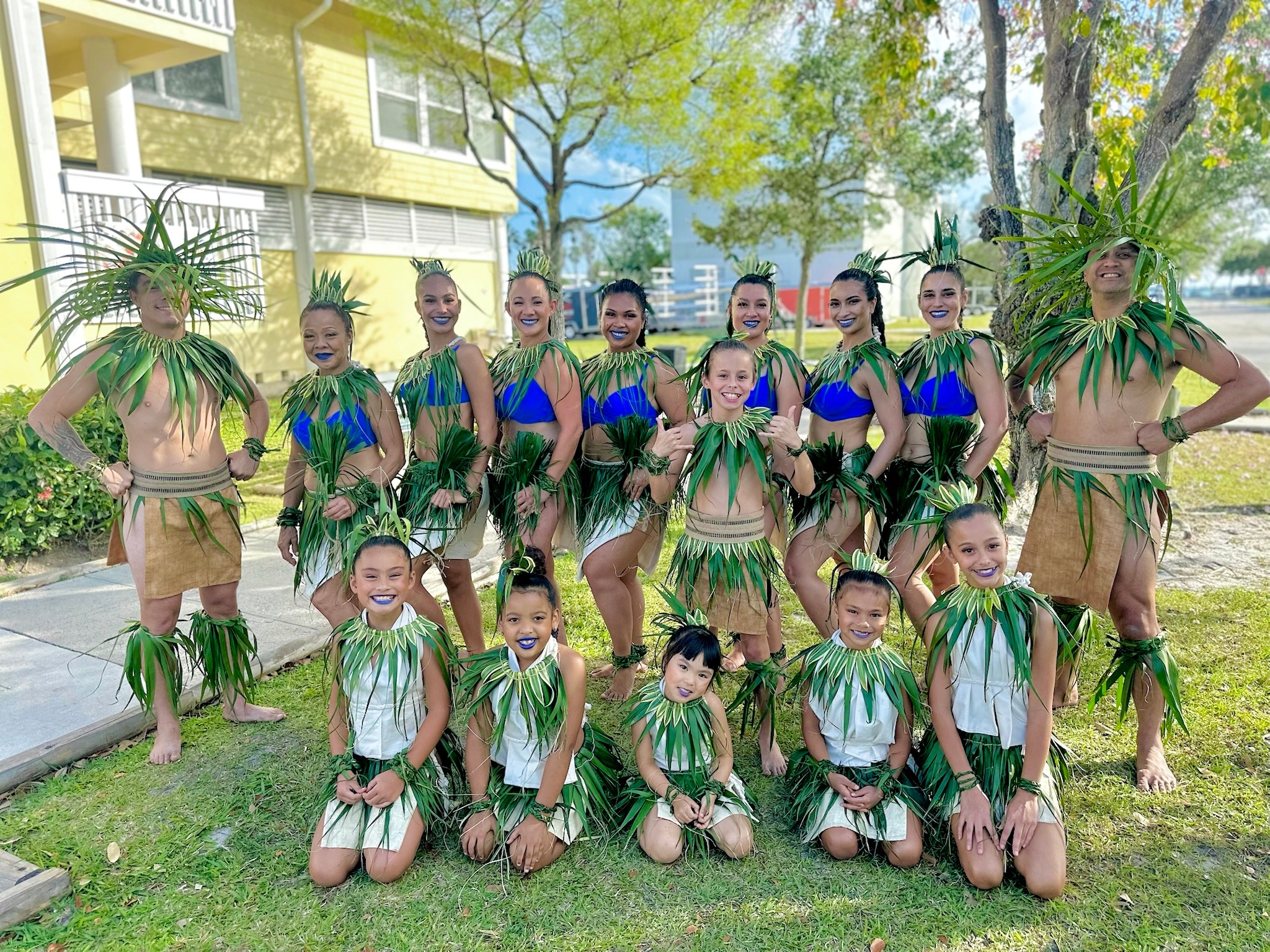 Group of children and adults in traditional costumes outdoors, with trees and buildings in background.