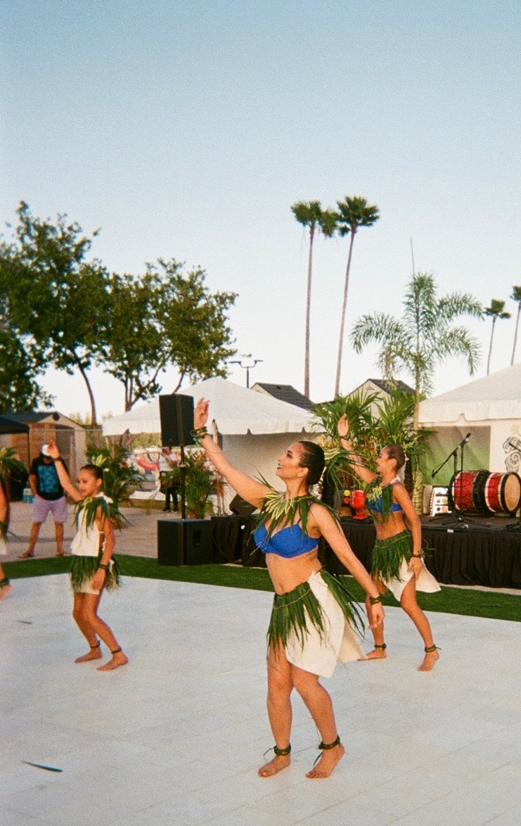 Group of women dancing outdoors with palm trees and tents in background, sunny sky, and musical instruments nearby.
