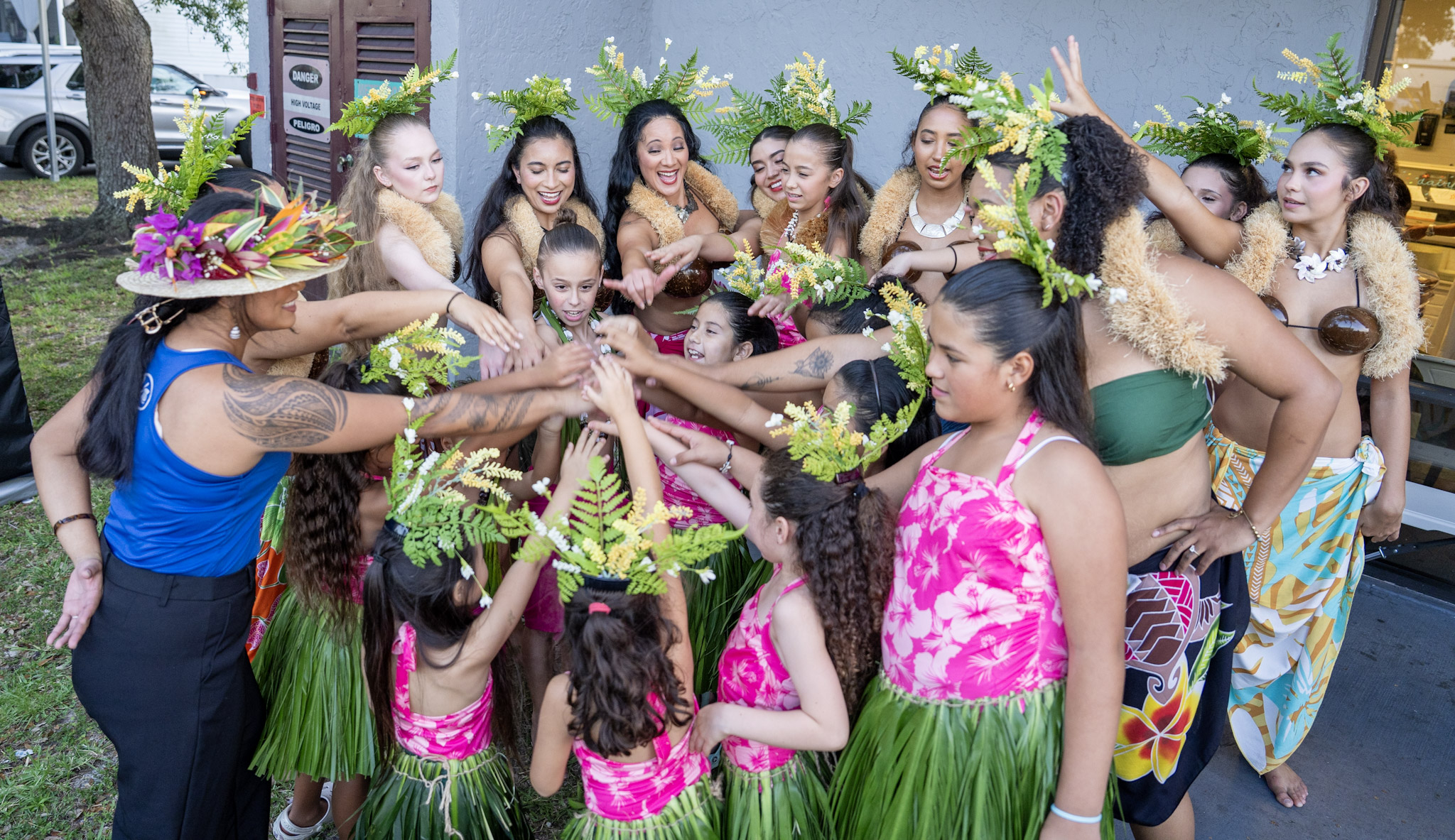 Group of women and children in traditional attire and floral headpieces, participating in a cultural dance outdoors.
