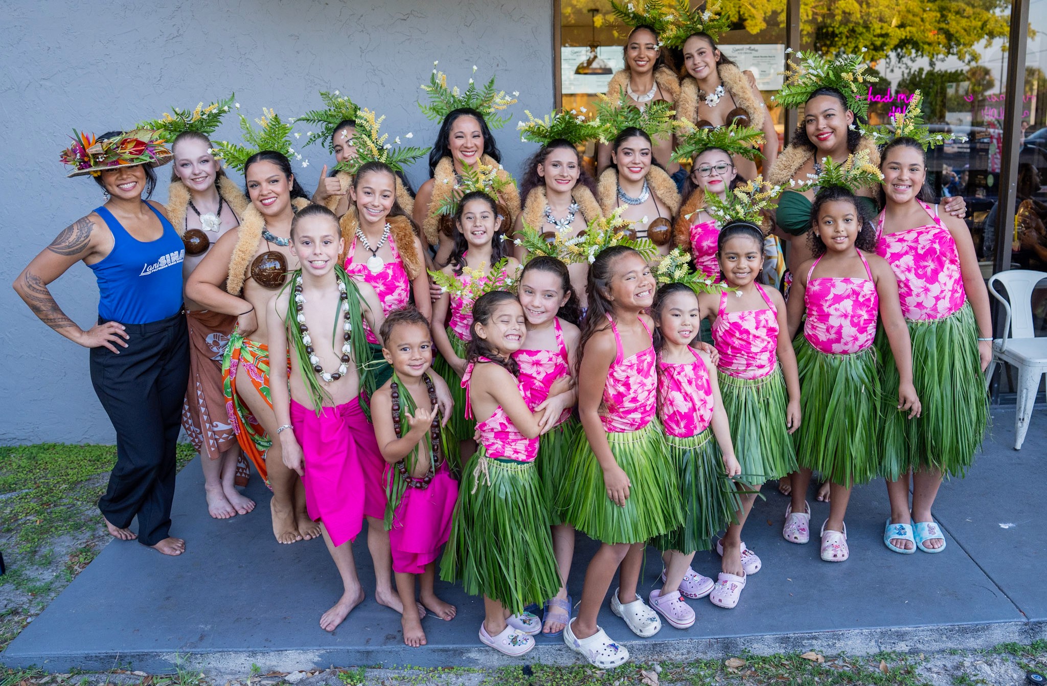 Group of young girls in colorful costumes and green skirts, posing outdoors with adults, flowers, and a building in background.