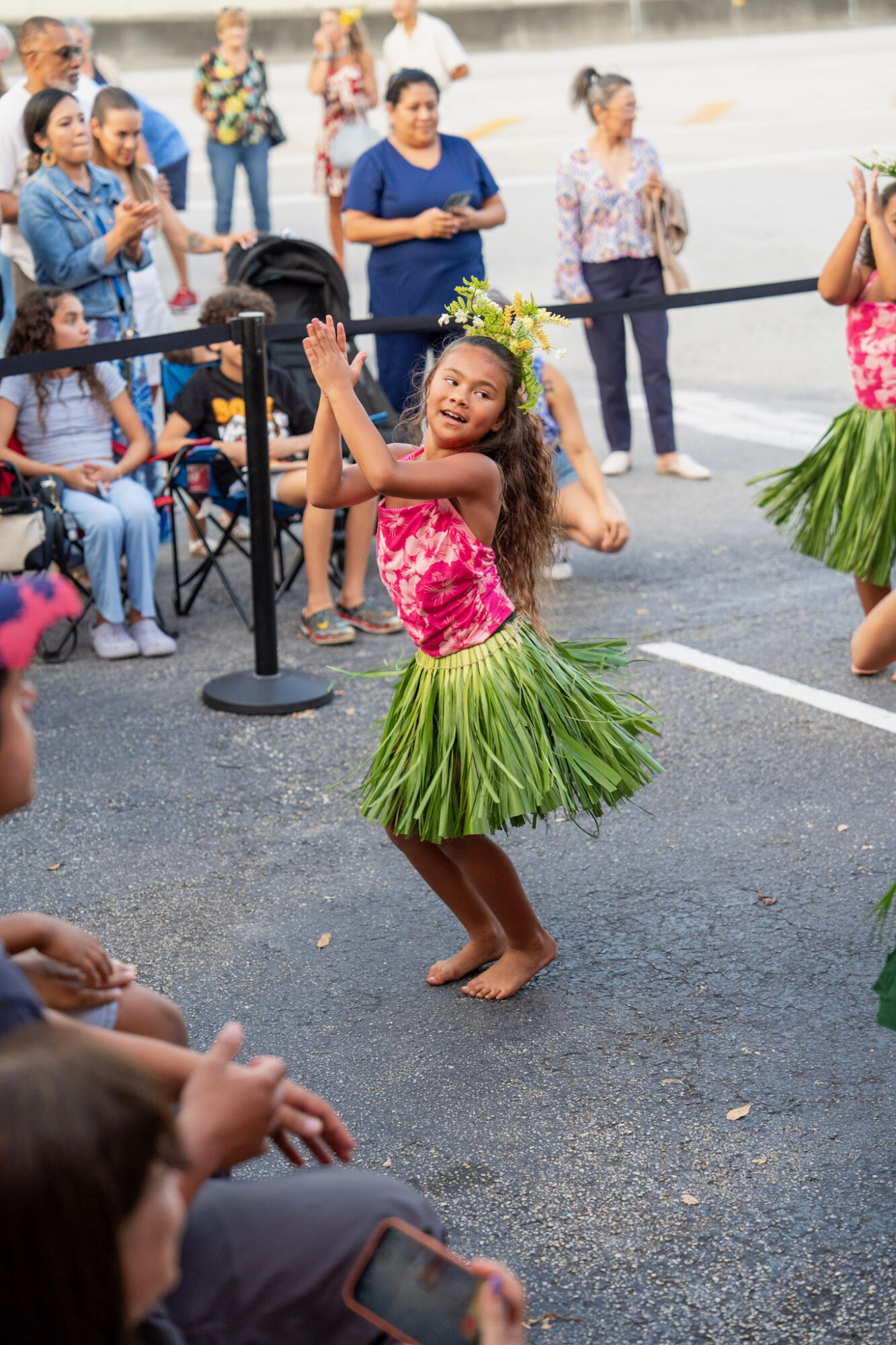 Young girl in a pink floral top and green grass skirt dancing outdoors, surrounded by onlookers, some taking photos.