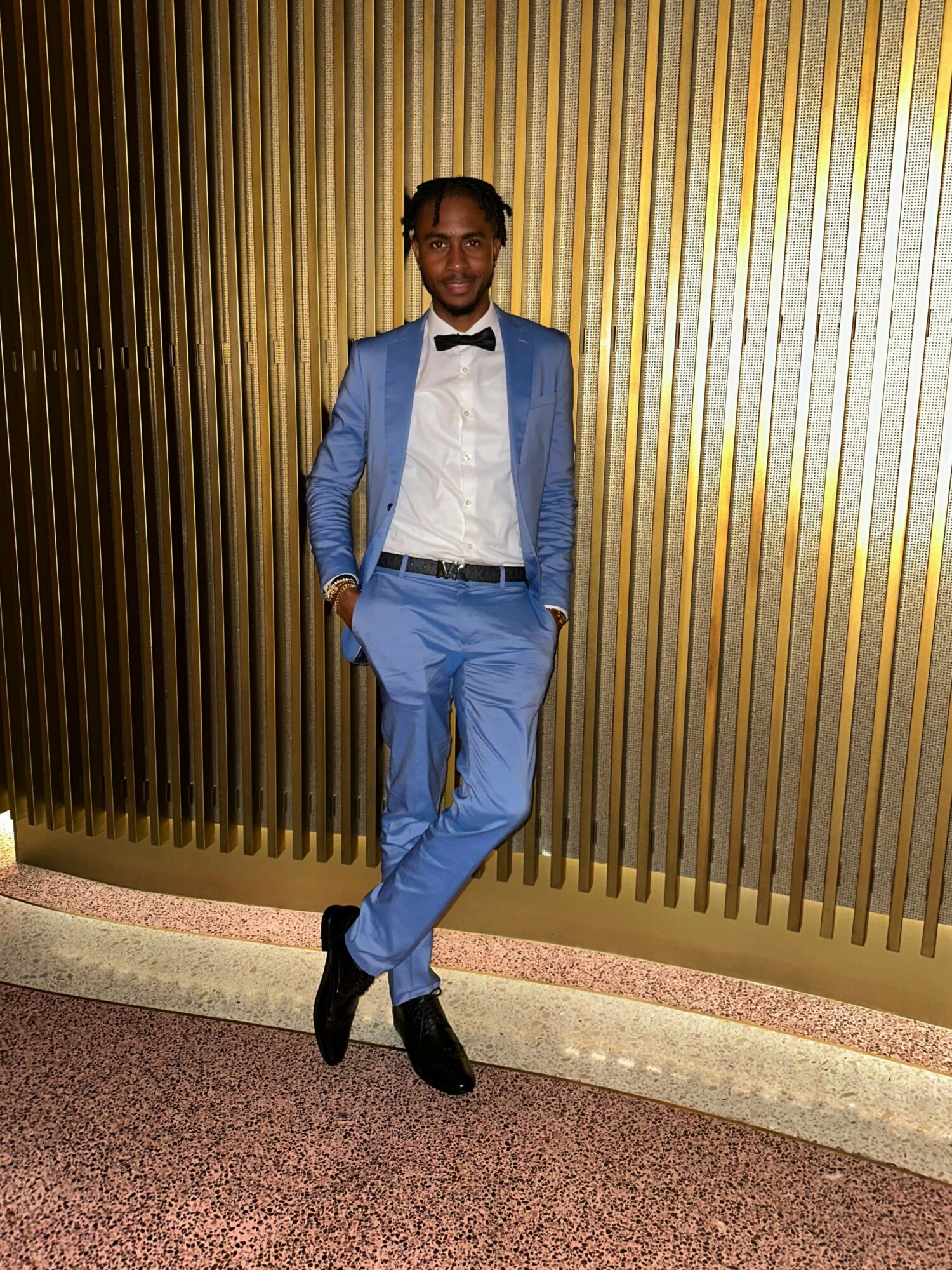 Man in a blue suit with a white shirt and black bow tie standing against a gold striped wall, hands in pockets.