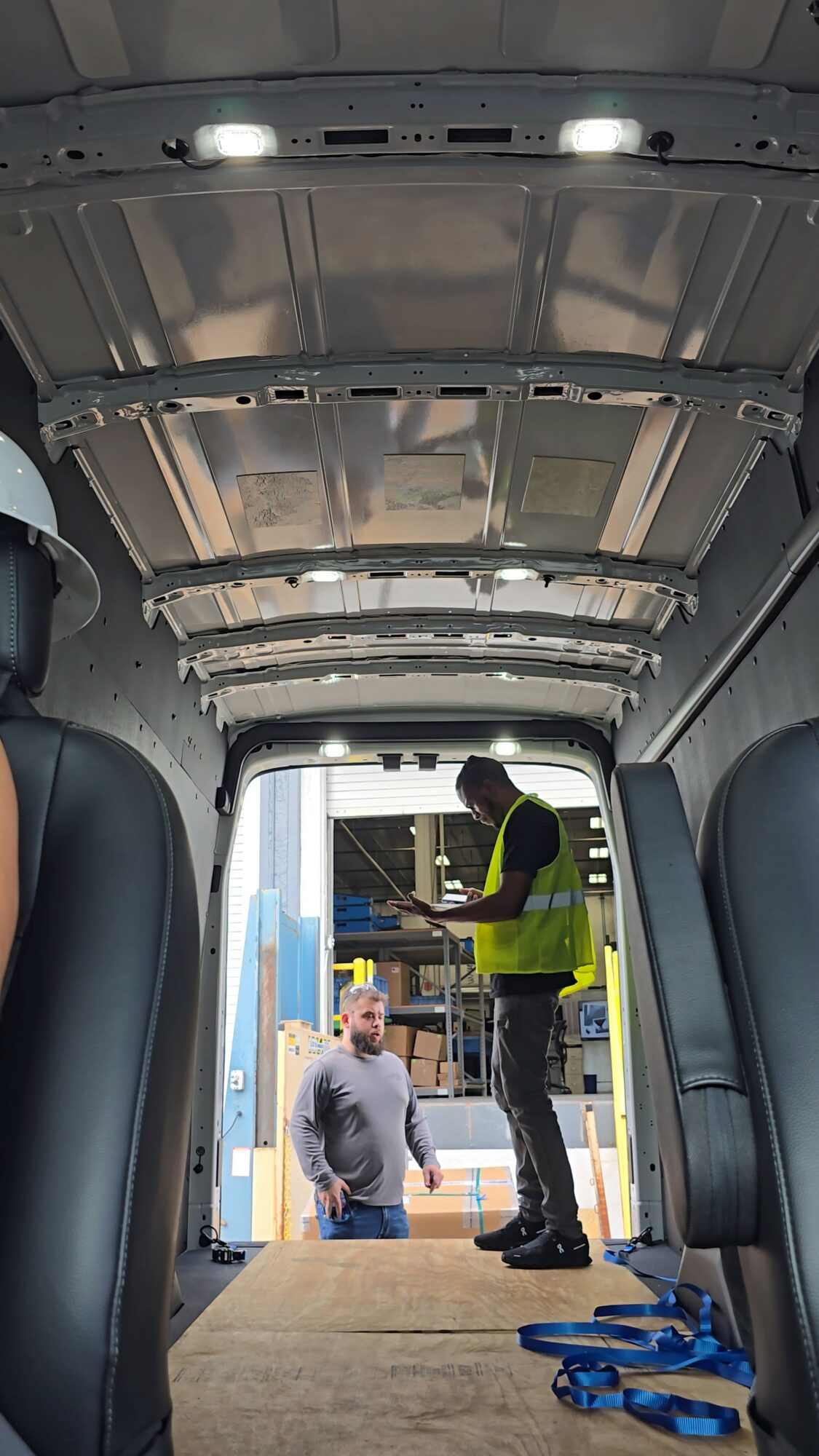 View from inside a vehicle looking out at two people, one in a high-visibility vest, near an open cargo door.