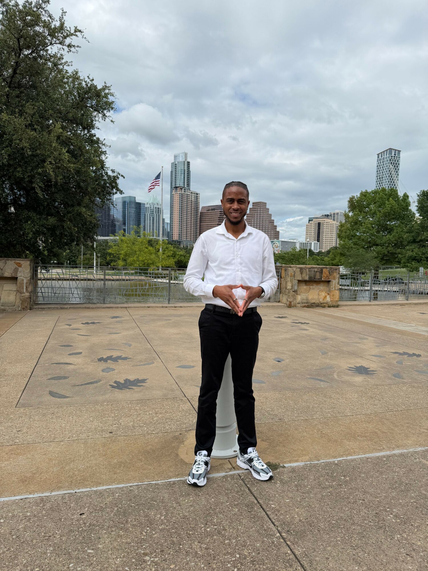 Man standing outdoors on concrete with city skyline in background, wearing white shirt and black pants.