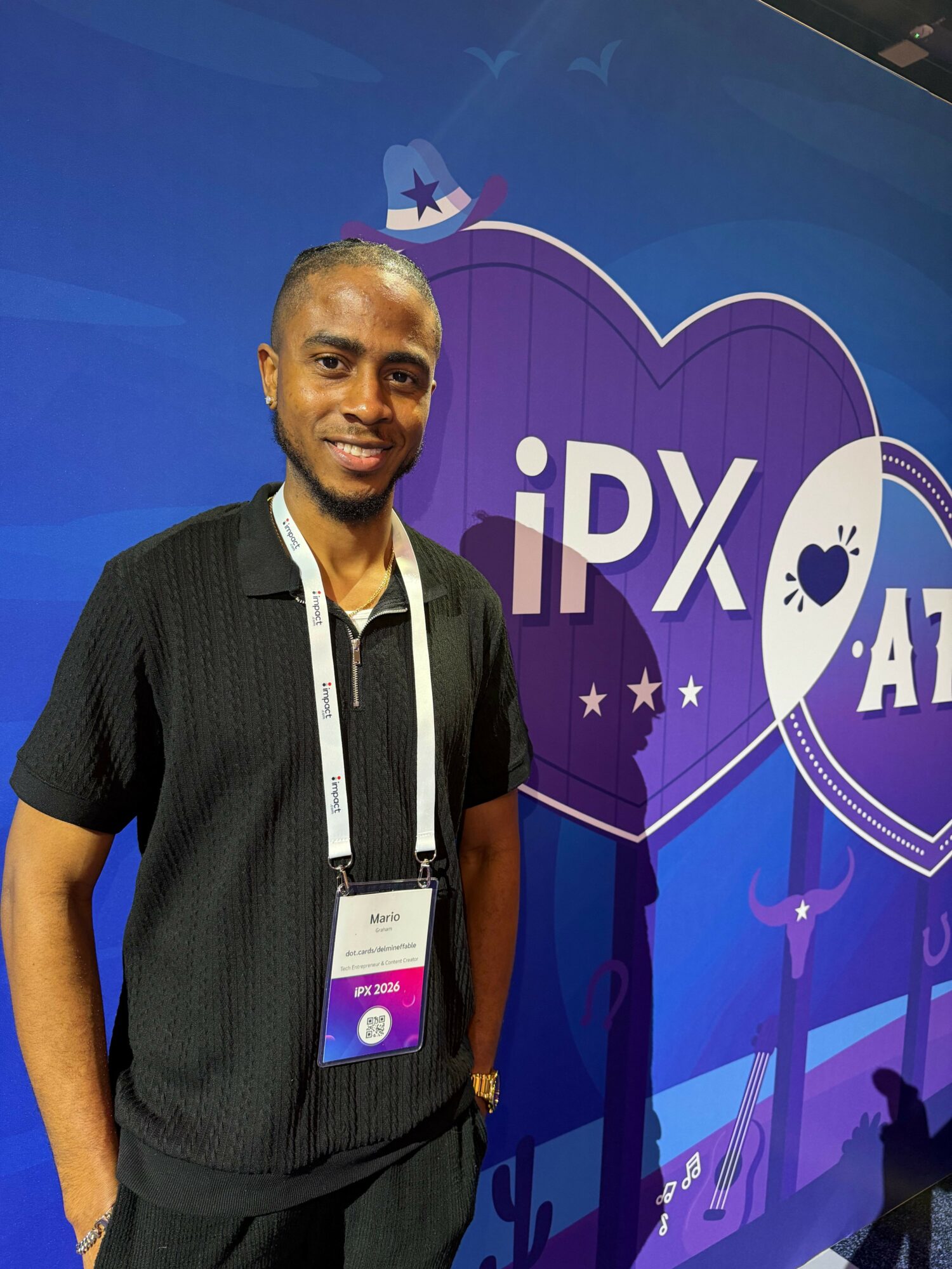 Man standing in front of a purple and blue event backdrop with text and graphics, smiling, wearing a black shirt and lanyard.