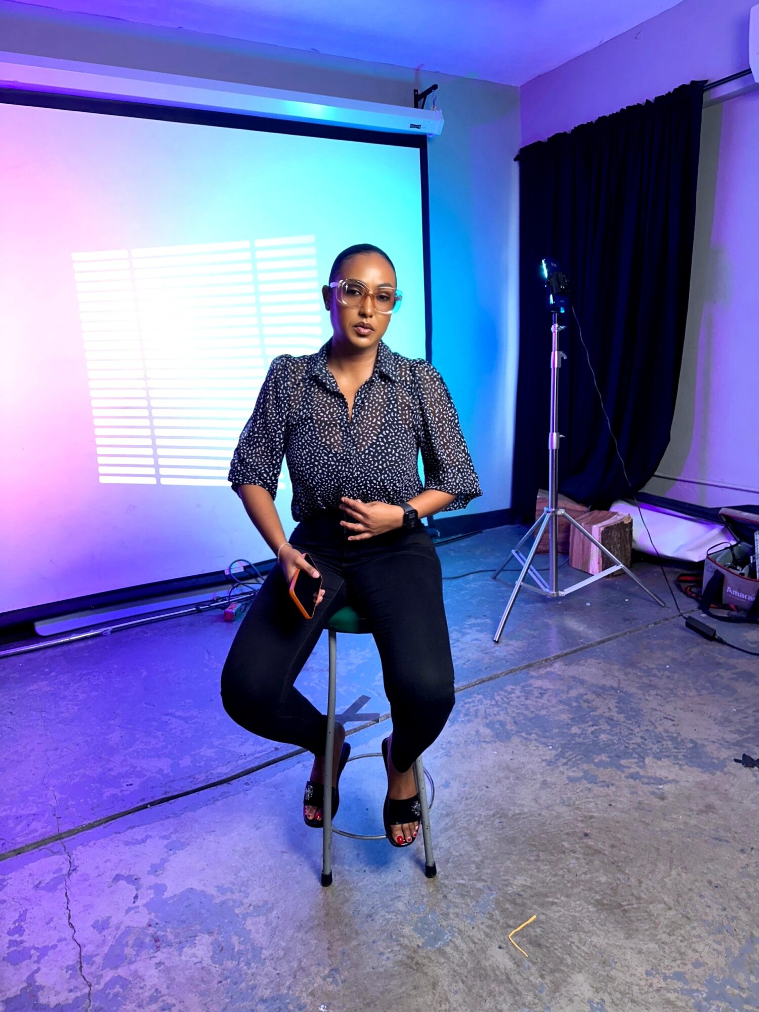 Woman sitting on a stool in a room with a projection screen and equipment, wearing glasses, a patterned blouse, and black pants.