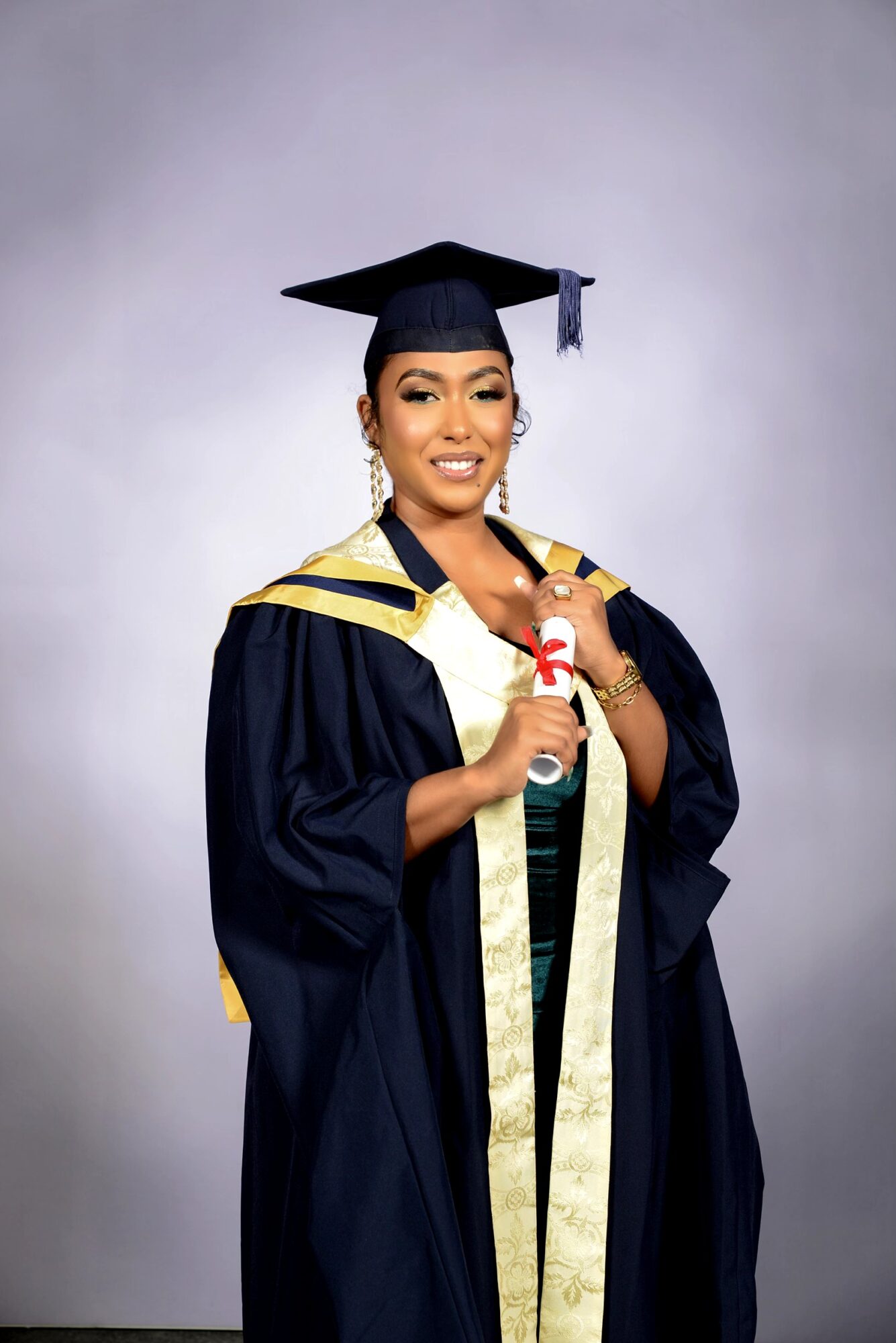 Woman in graduation gown and cap holding diploma, smiling, against a plain background.