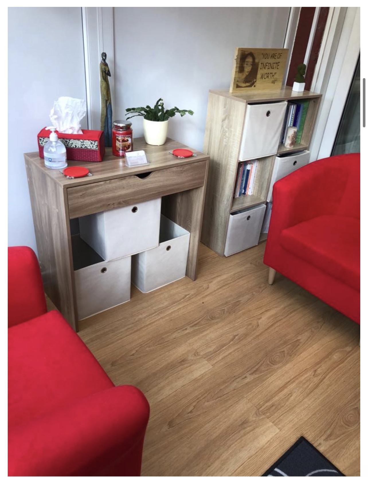 Room corner with wooden furniture, red sofa, potted plant, books, and decorative items, wooden floor, and a window with curtains.