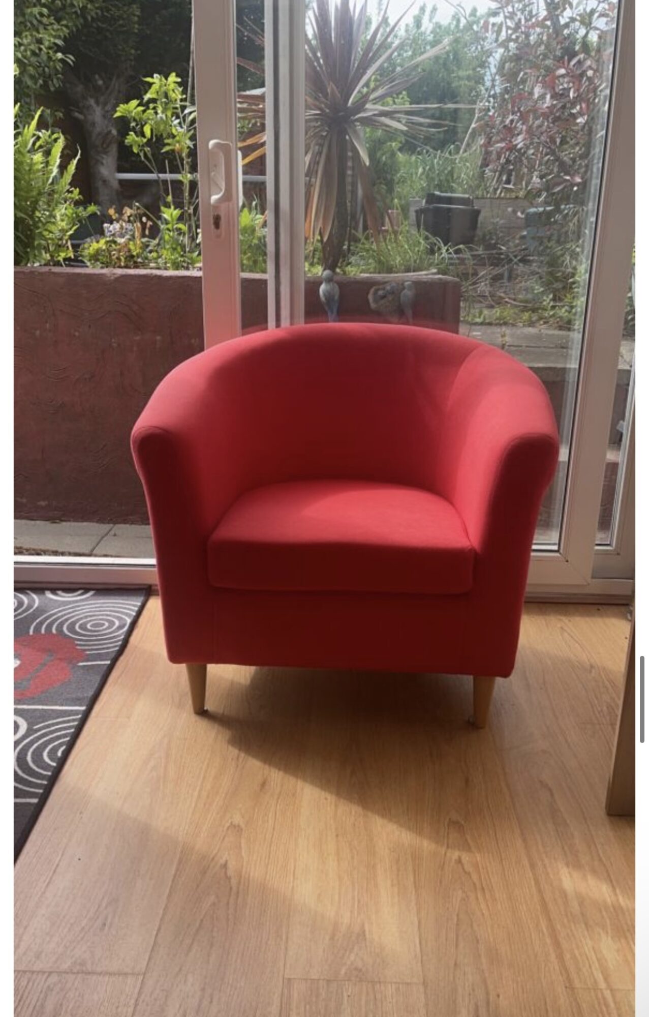 Red armchair on wooden floor in front of glass door with garden view, sunlight casting shadows.