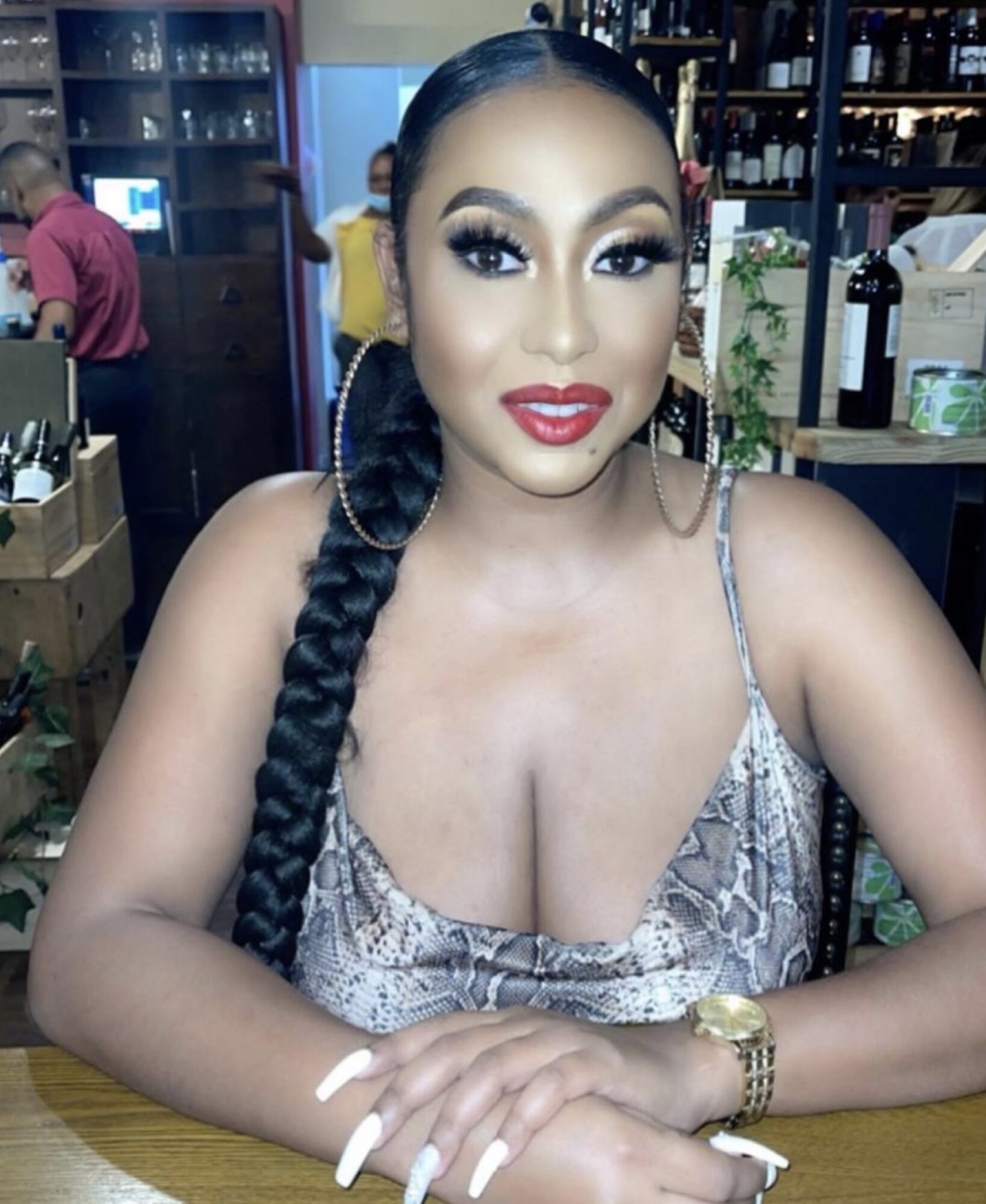 Woman with long braided hair, makeup, and a patterned top sitting at a table in a bar or restaurant.
