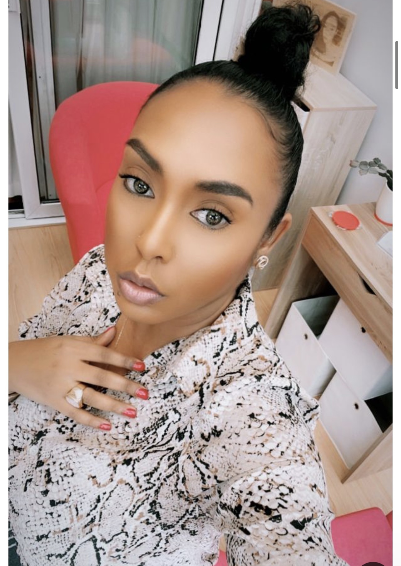 Woman with dark hair in a high bun, wearing a snake print top, sitting in a room with furniture and a red chair.