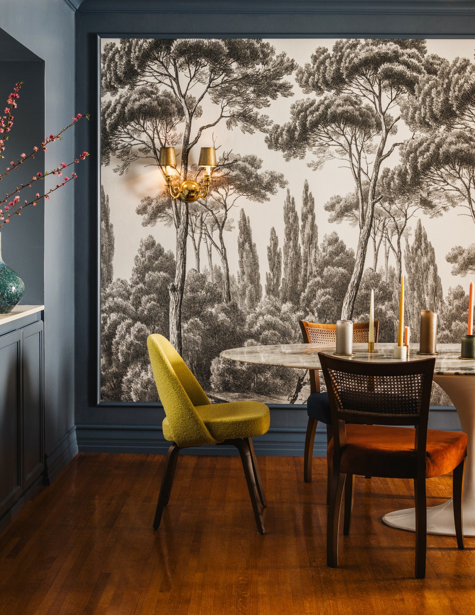 Dining room with a large forest mural, yellow and black chairs, a marble table, and candles, with a wall sconce and vase.