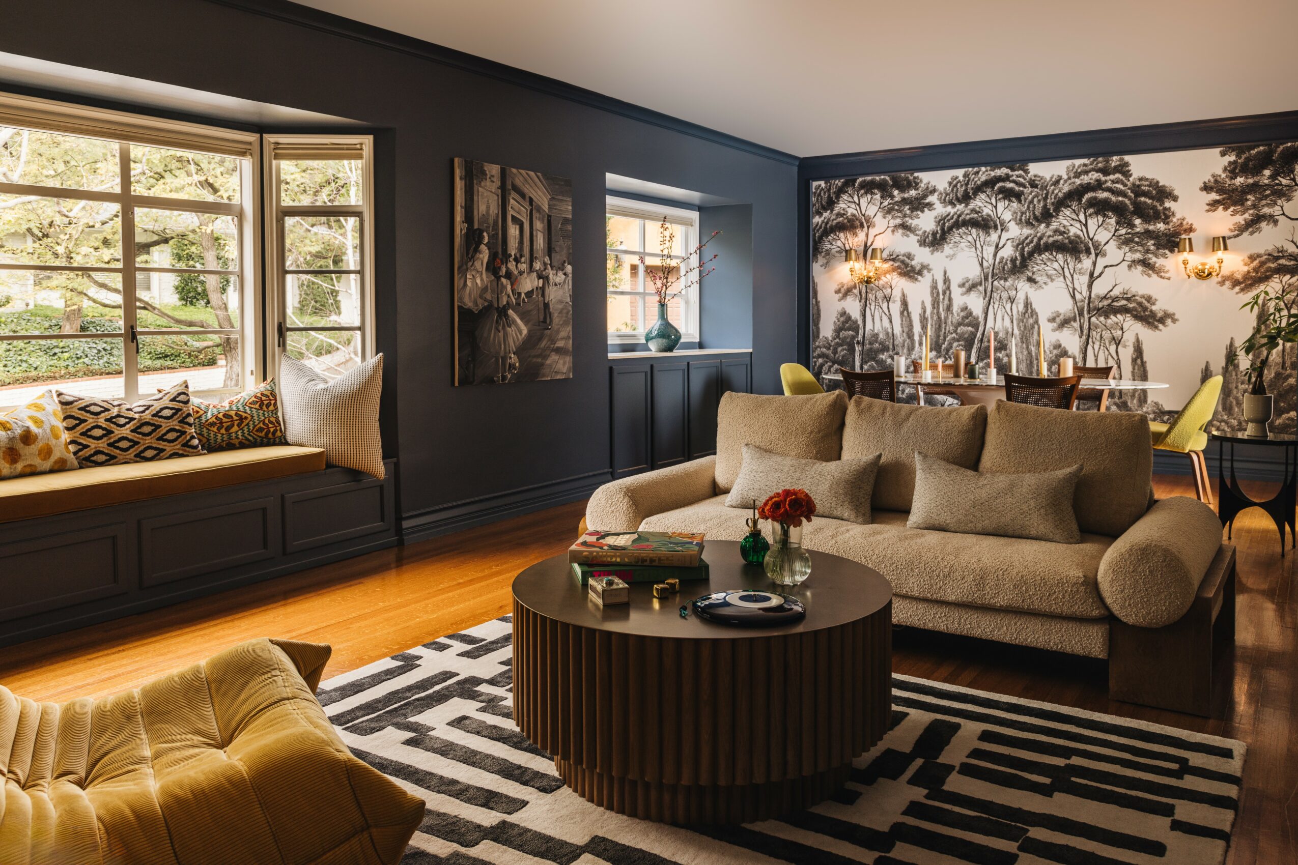 Living room with large window, beige sofa, black and white rug, round coffee table, and wall mural of trees.