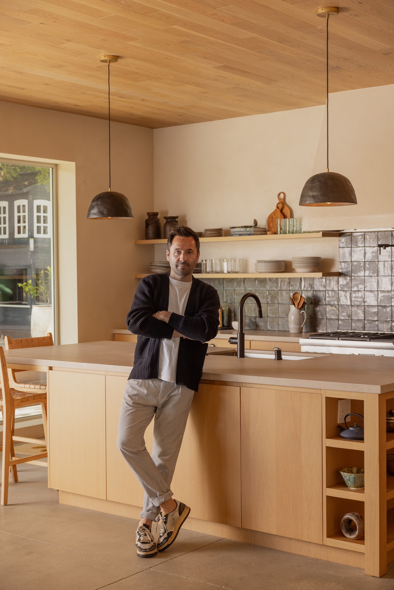 Man standing in a modern kitchen with wooden cabinets, hanging pendant lights, and a window, leaning against the counter.