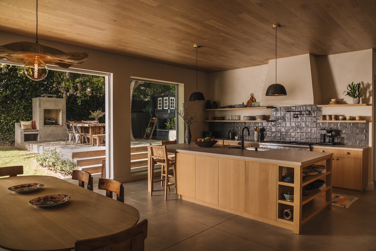 Modern kitchen with wooden island, open shelves, and large window showing outdoor patio and greenery.