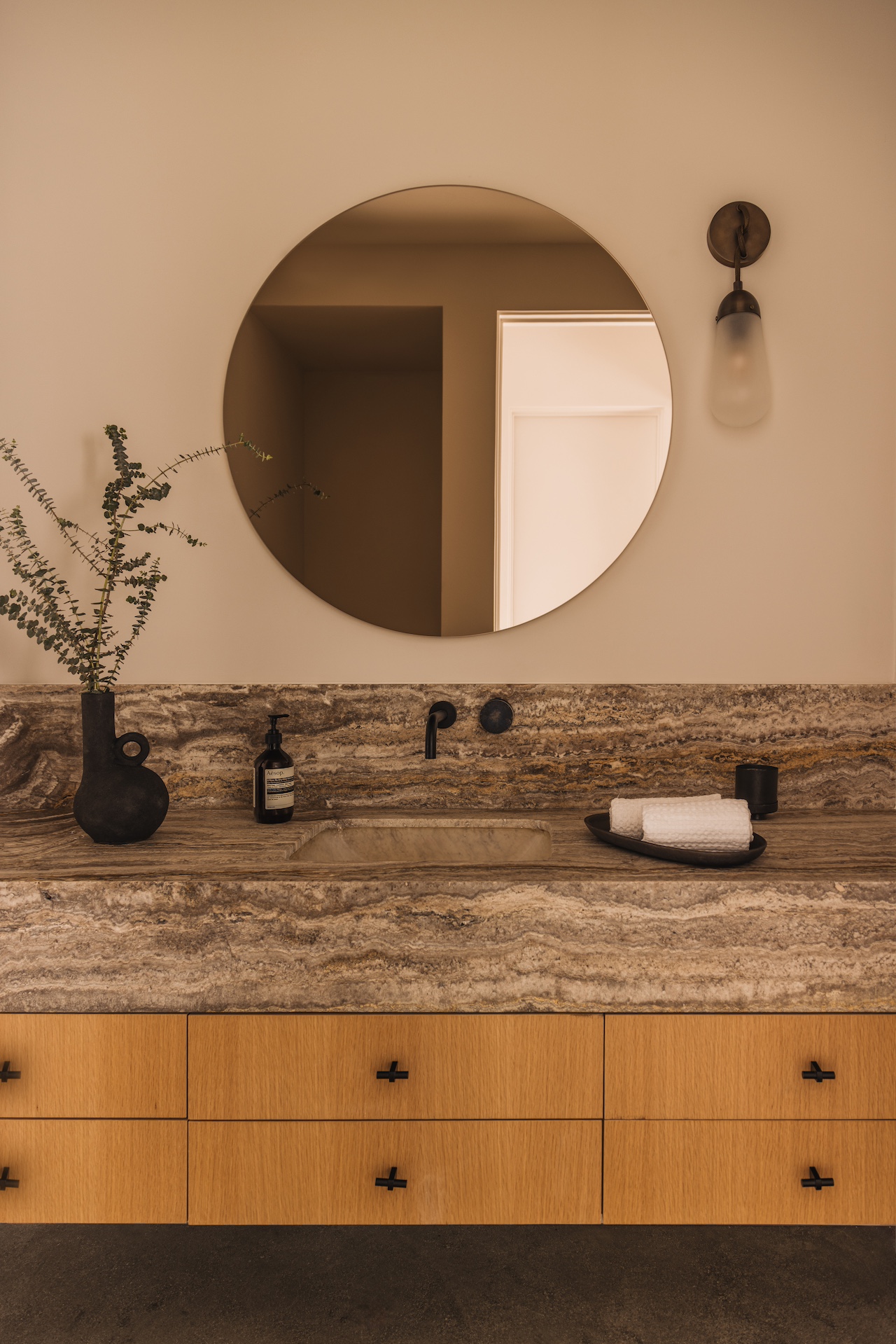 Bathroom vanity with a round mirror, a black vase with branches, soap dispenser, and rolled towel on a wooden countertop.