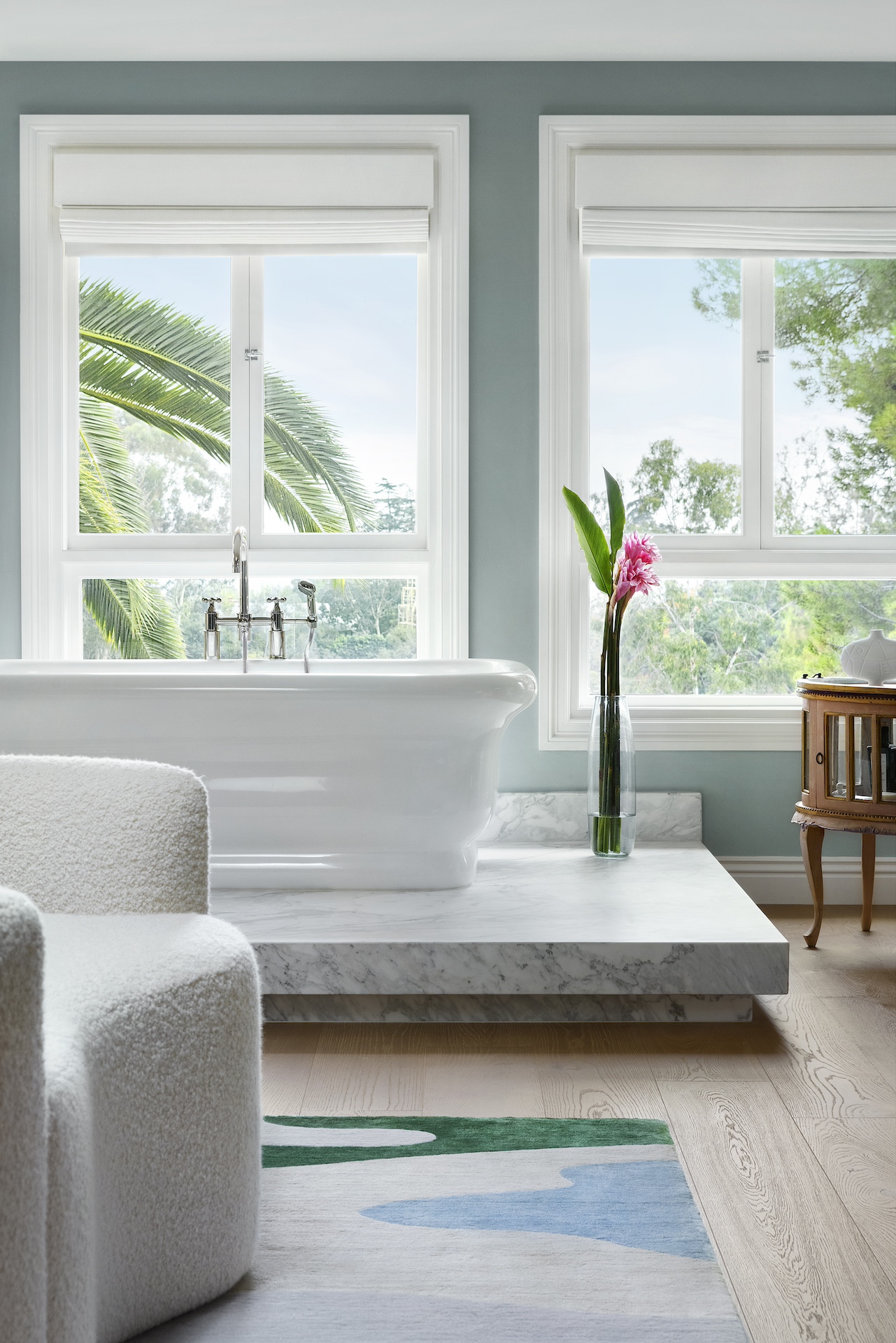 Bathroom with a bathtub, large windows, a tall vase with pink flowers, and a wooden chair, bright and airy.