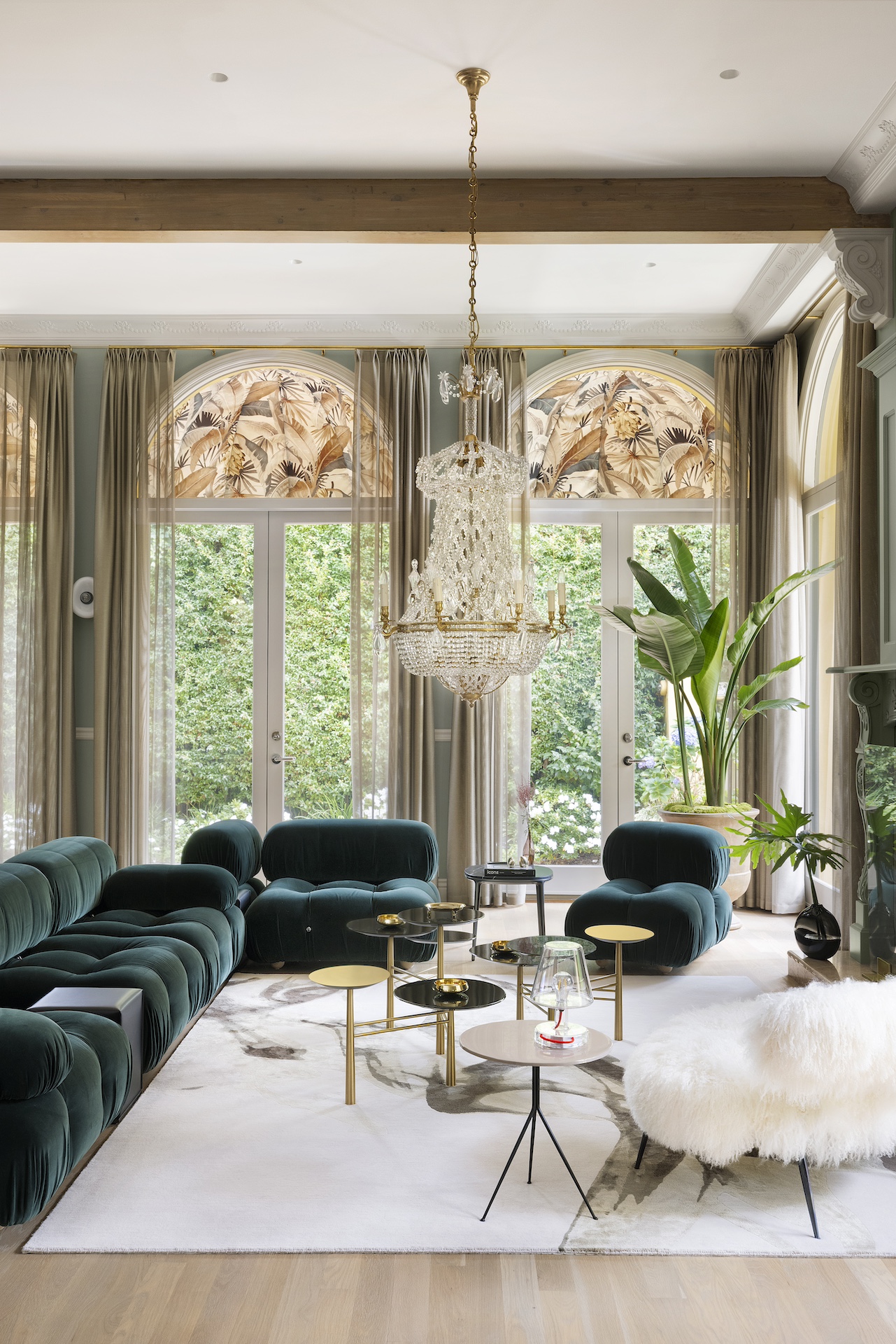 Elegant living room with large windows, velvet sofas, a chandelier, and a tall plant, brightened by natural light.