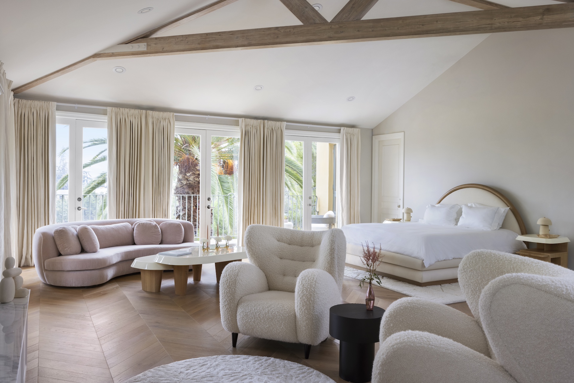 Bright bedroom with large windows, pink sofa, white armchairs, and a bed with a curved headboard, wooden floor, and ceiling beams.