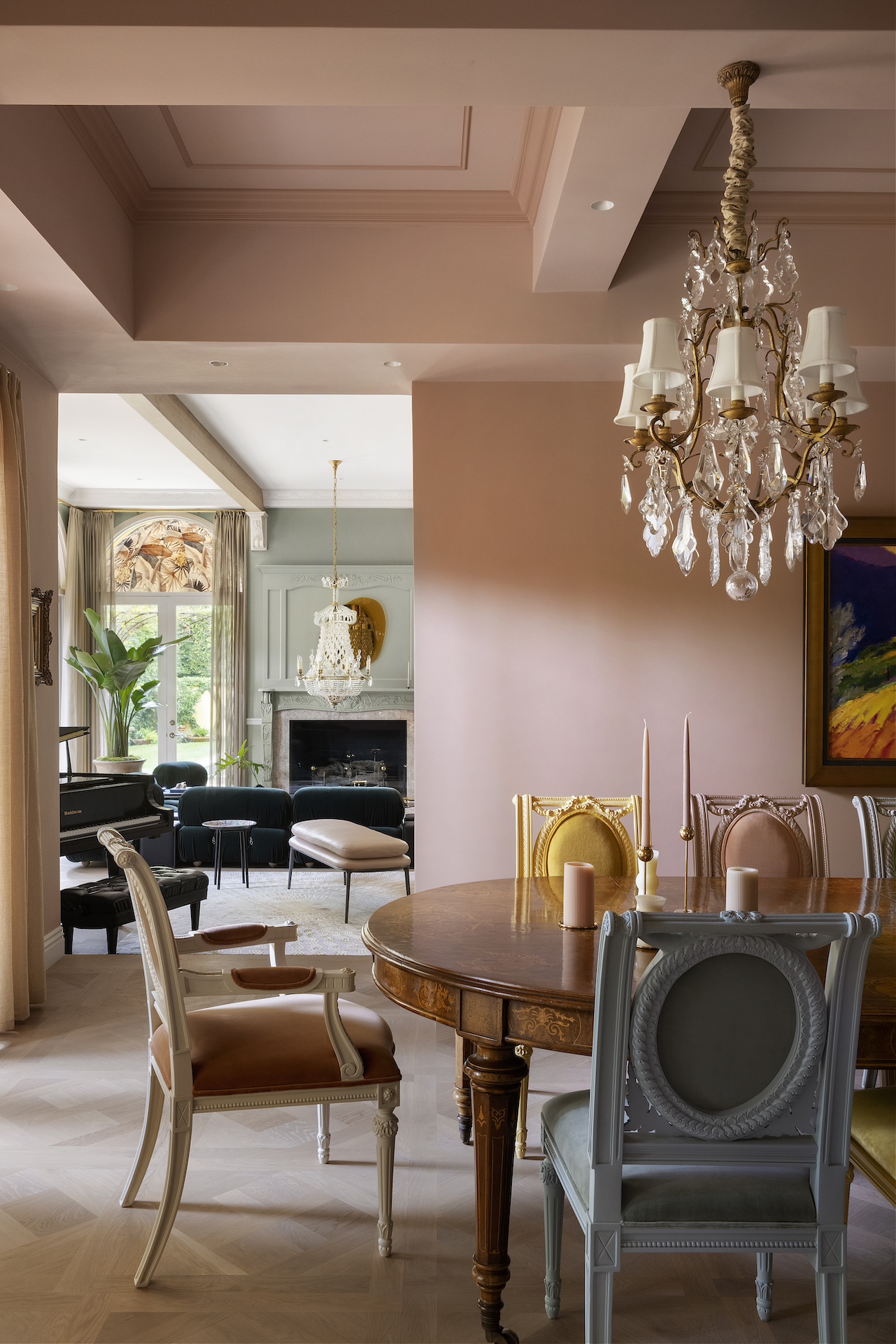 Elegant dining room with chandelier, round wooden table, and chairs, with a living area and large window in the background.