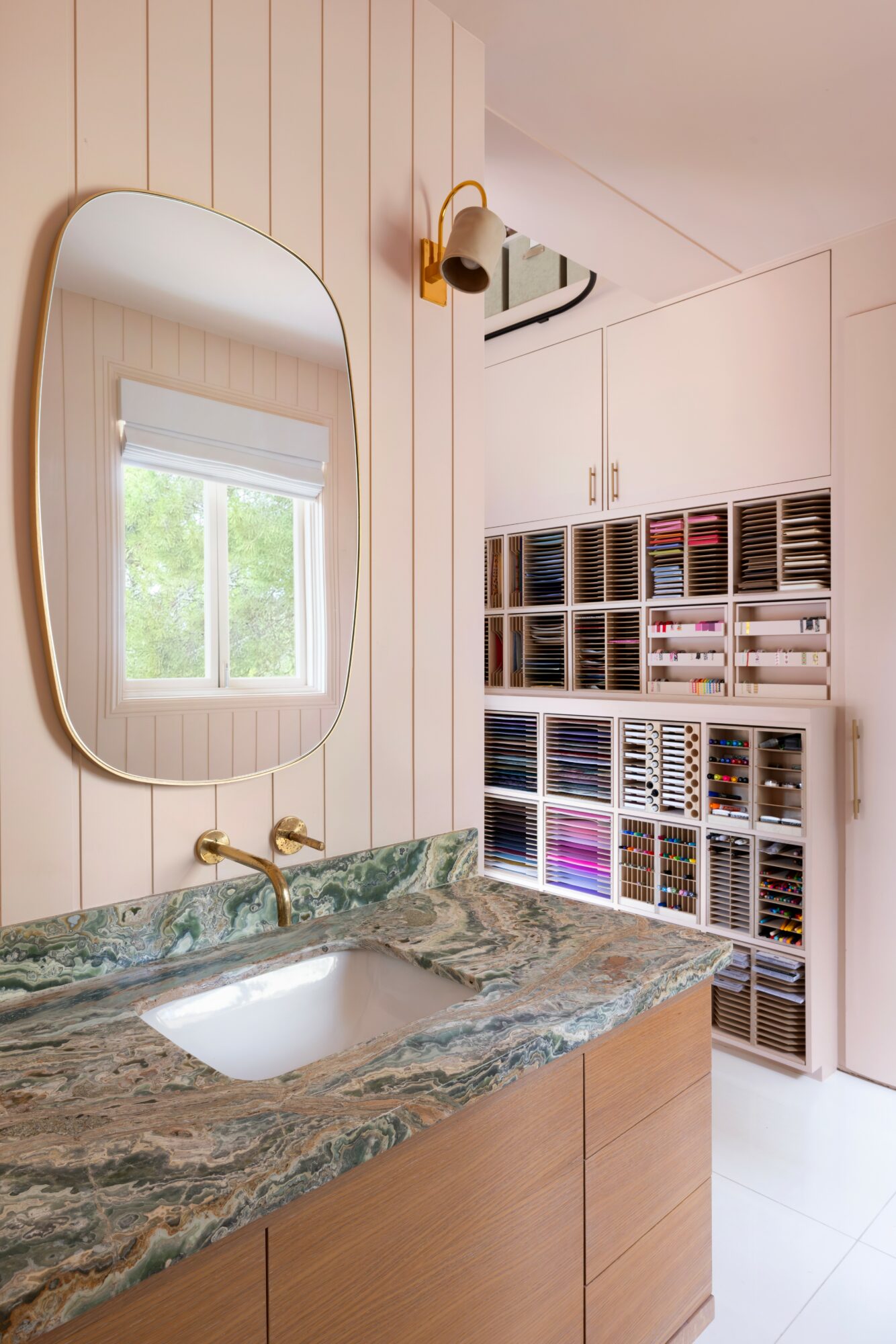 Bathroom vanity with a marble countertop, oval mirror, window, and storage shelves with organized items.