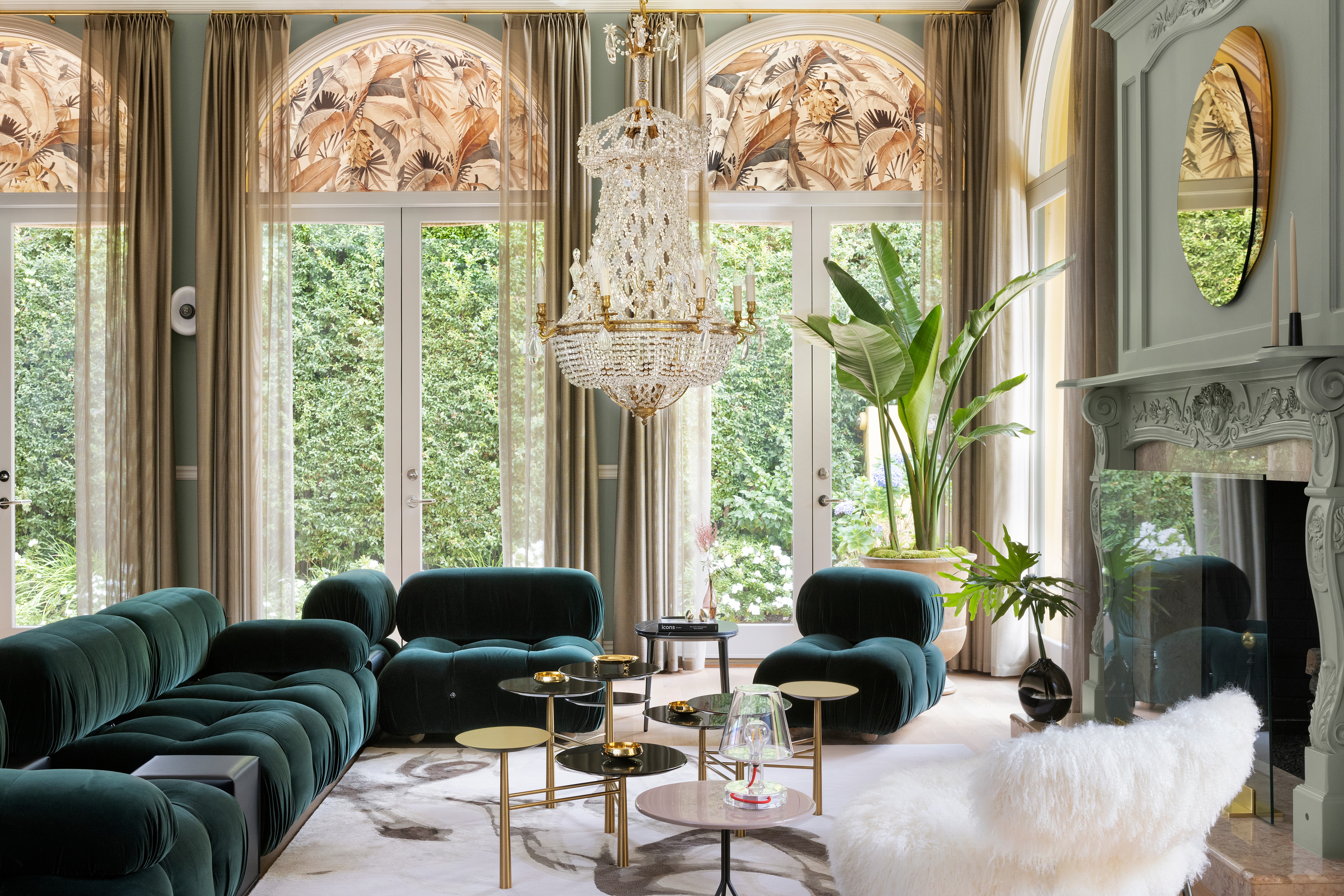 Elegant living room with large windows, chandelier, green velvet sofas, and a fireplace, decorated with plants and curtains.