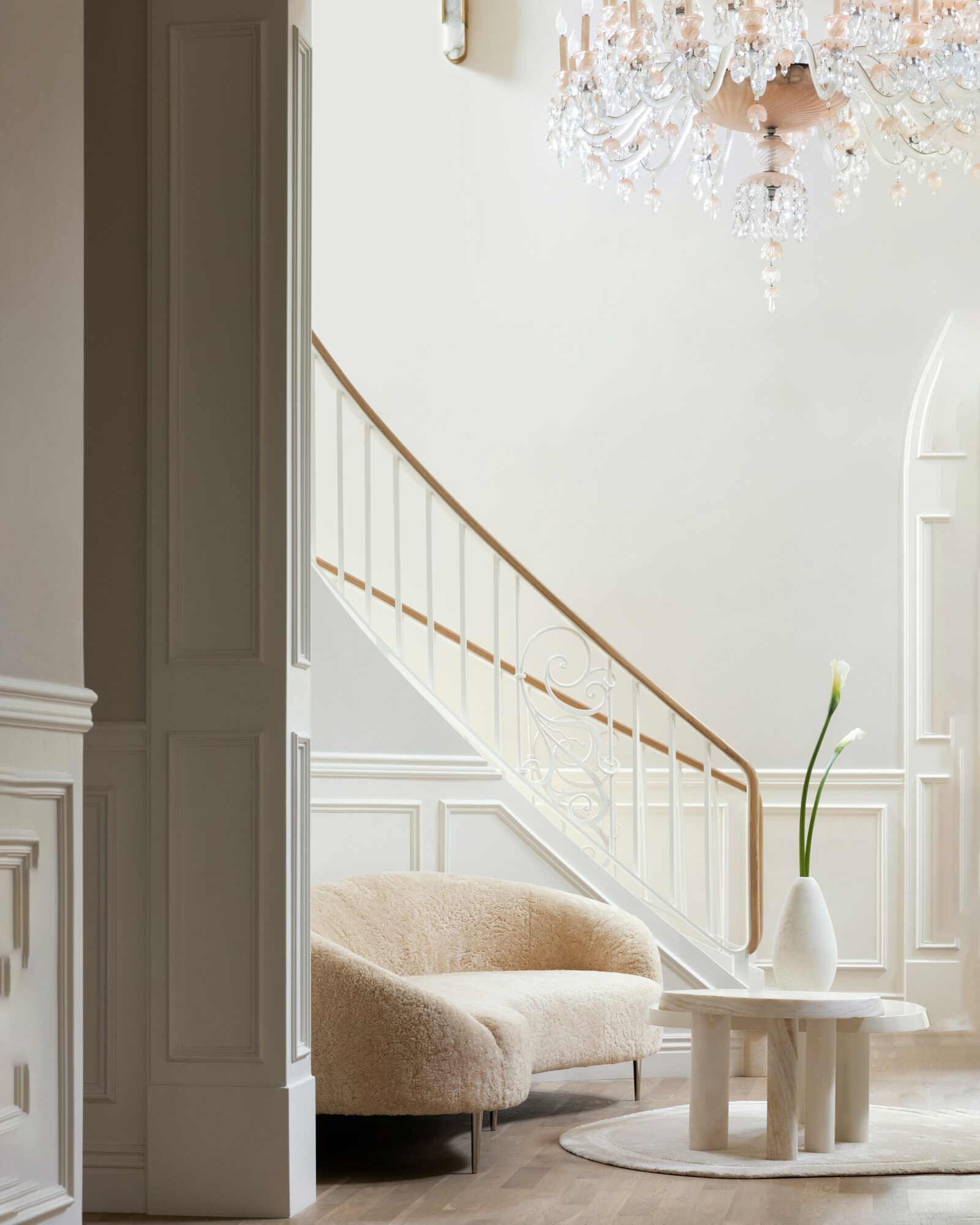 Elegant interior with a staircase, chandelier, beige sofa, and a white vase with a flower on a round table.