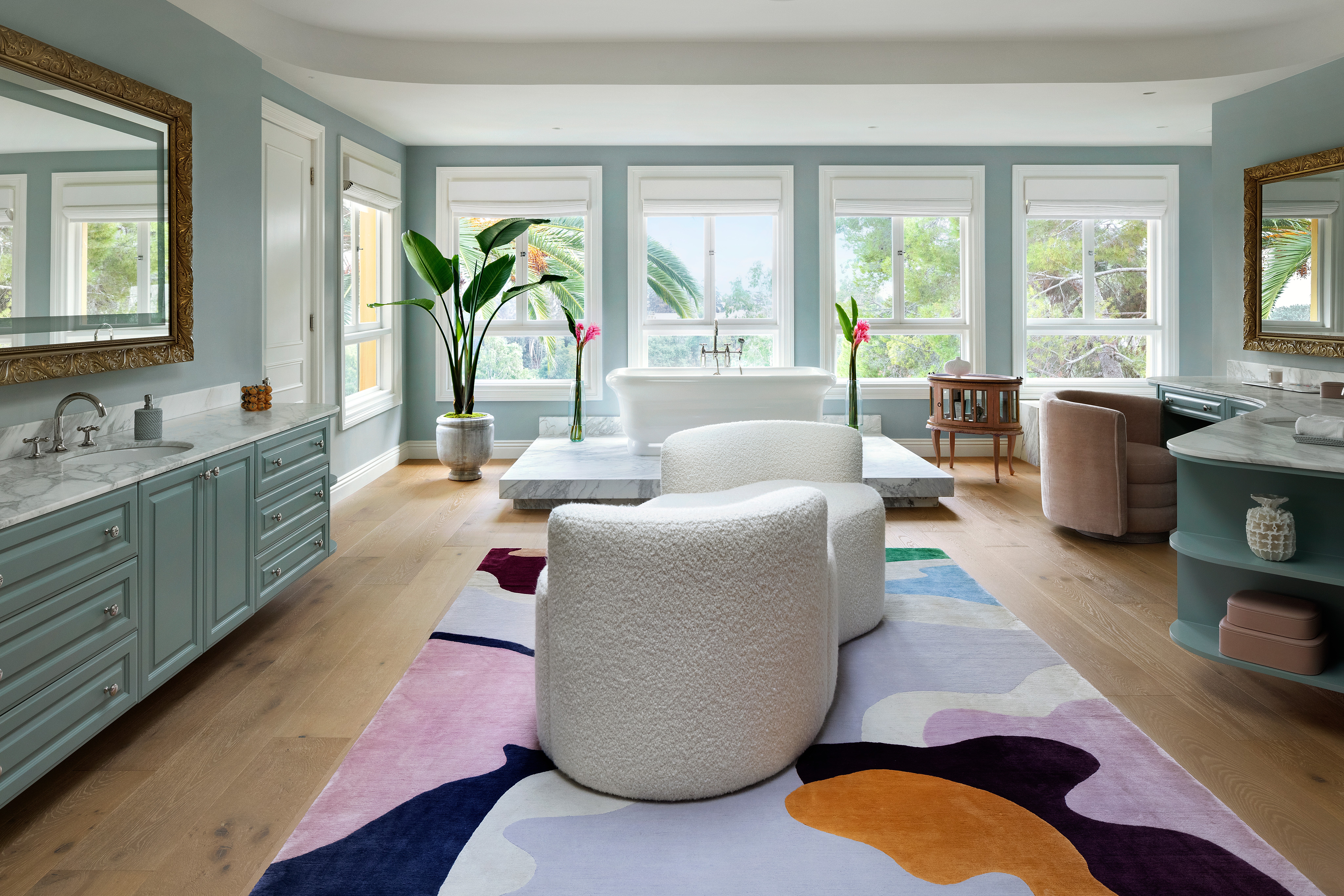 Bright living room with large windows, white furniture, colorful rug, and potted plants, decorated in pastel tones.