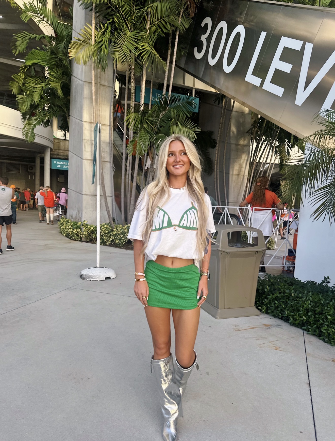 Woman with long blonde hair wearing a white crop top and green skirt, standing outdoors near a sign that reads '300 LEVEL'.