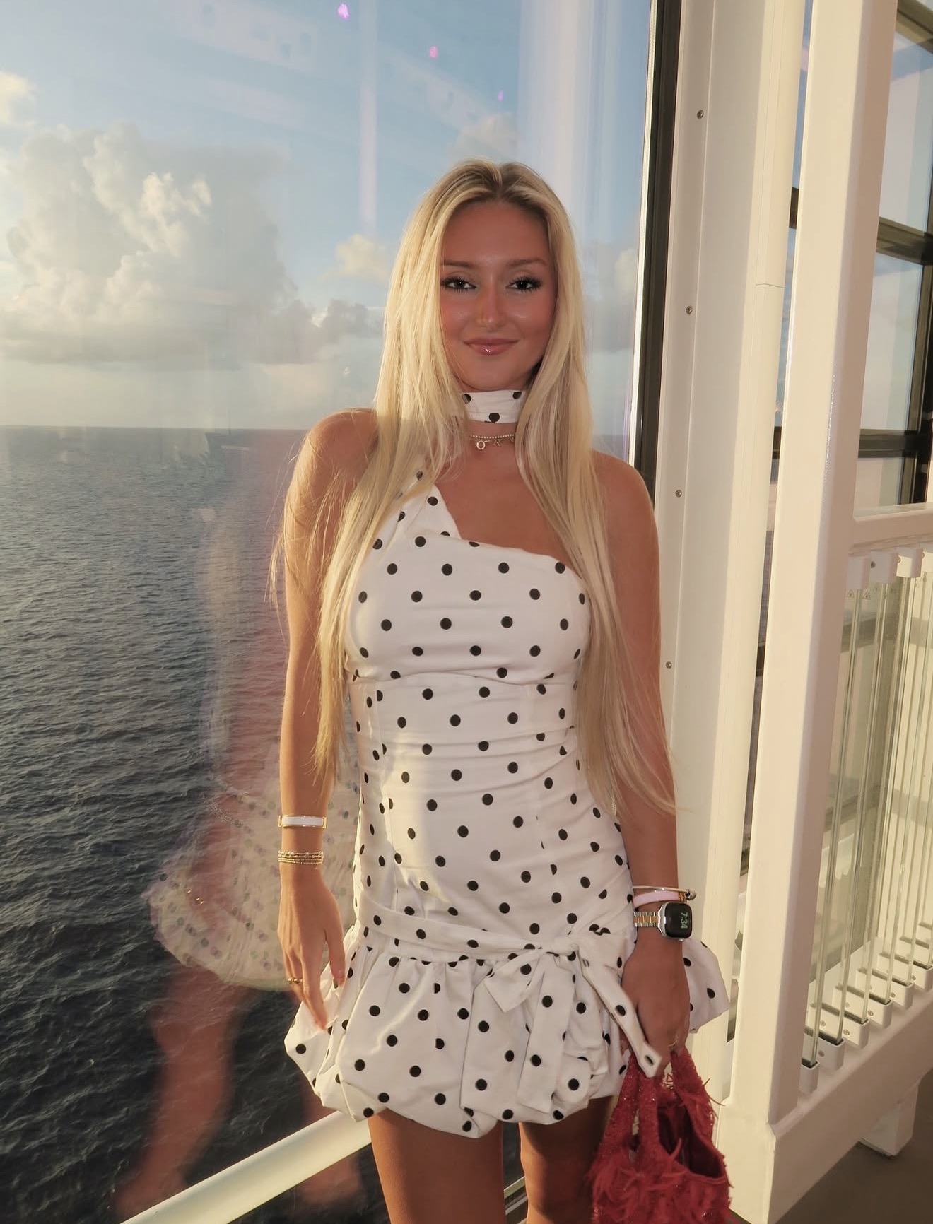 Young woman with long blonde hair in a polka dot dress standing by a window with ocean view, smiling.
