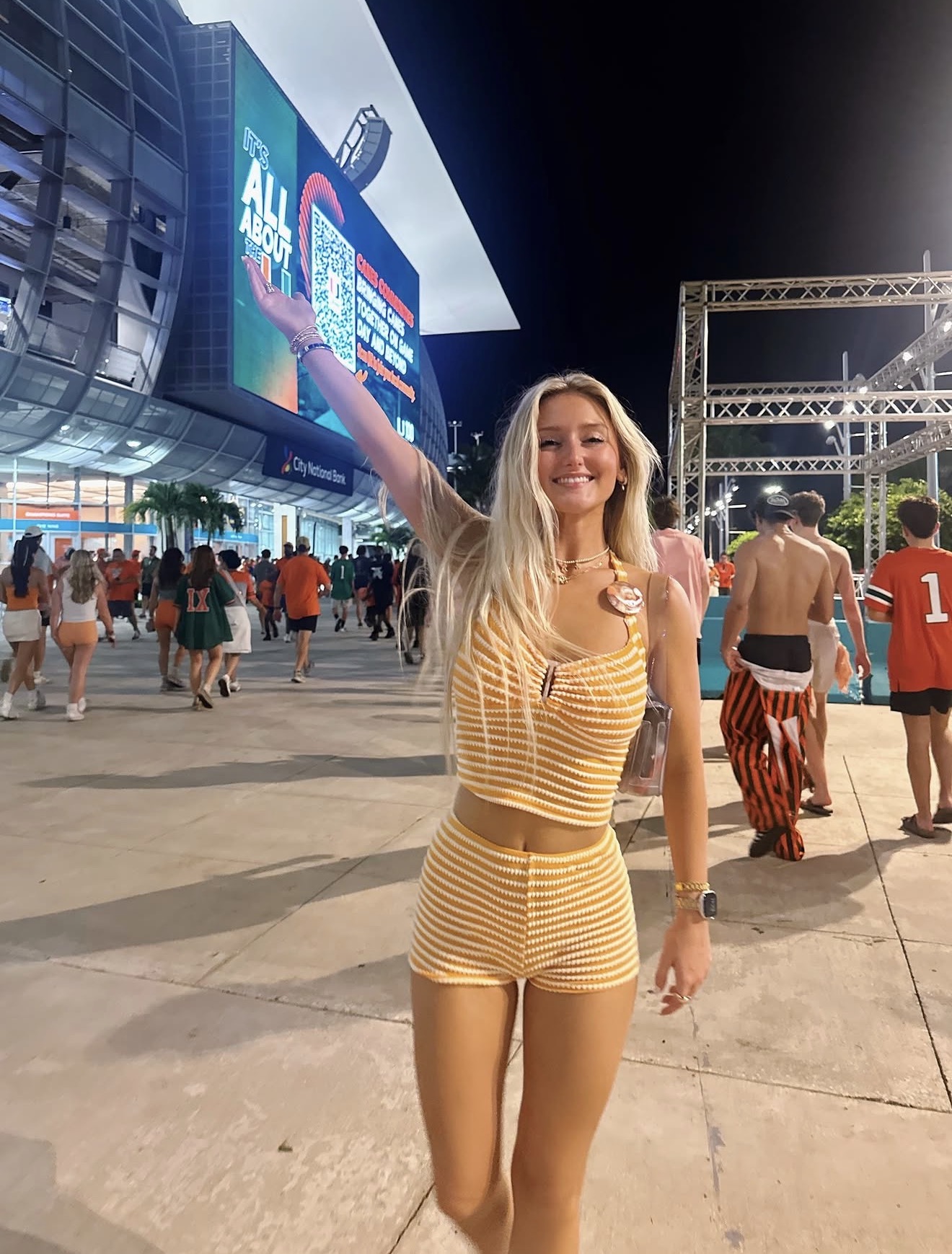 Smiling woman in yellow striped outfit waving at outdoor event with crowd and large screen in background.