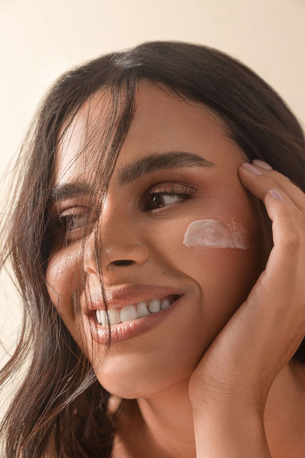 Smiling woman with long dark hair and a bandage on her cheek, touching her face gently.